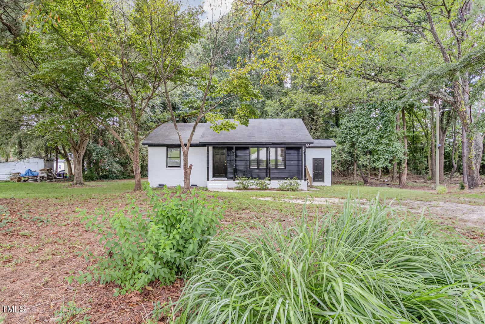 A small, single-story house with white walls and dark trim sits surrounded by trees and greenery at 302 Tar River Avenue in Louisburg. The front yard features shrubs and a mulch bed, with a wooded area in the background.