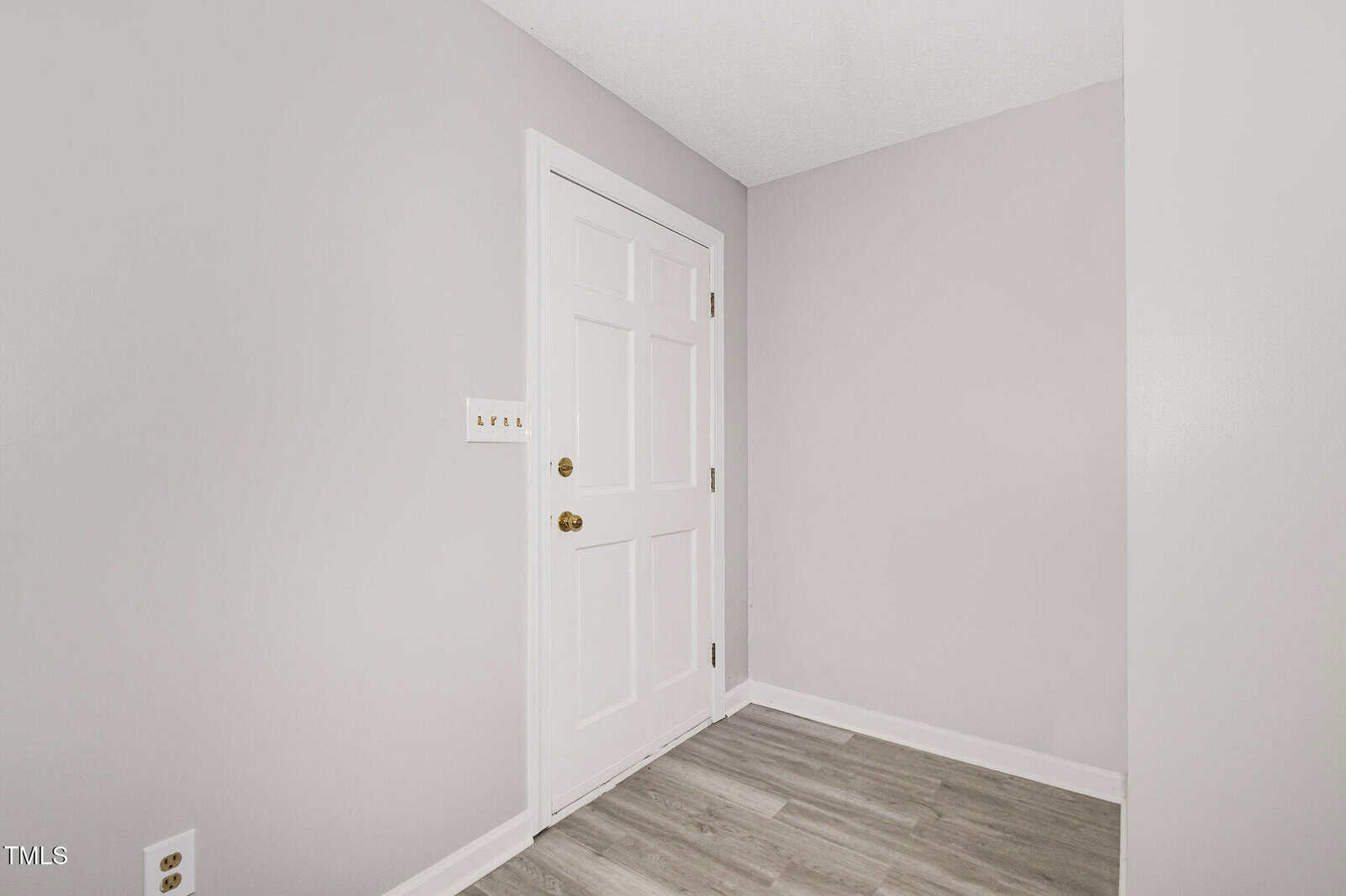 A small entryway with light gray walls, white trim, a white door with gold hardware, light wood flooring, and a three-toggle light switch on the wall.