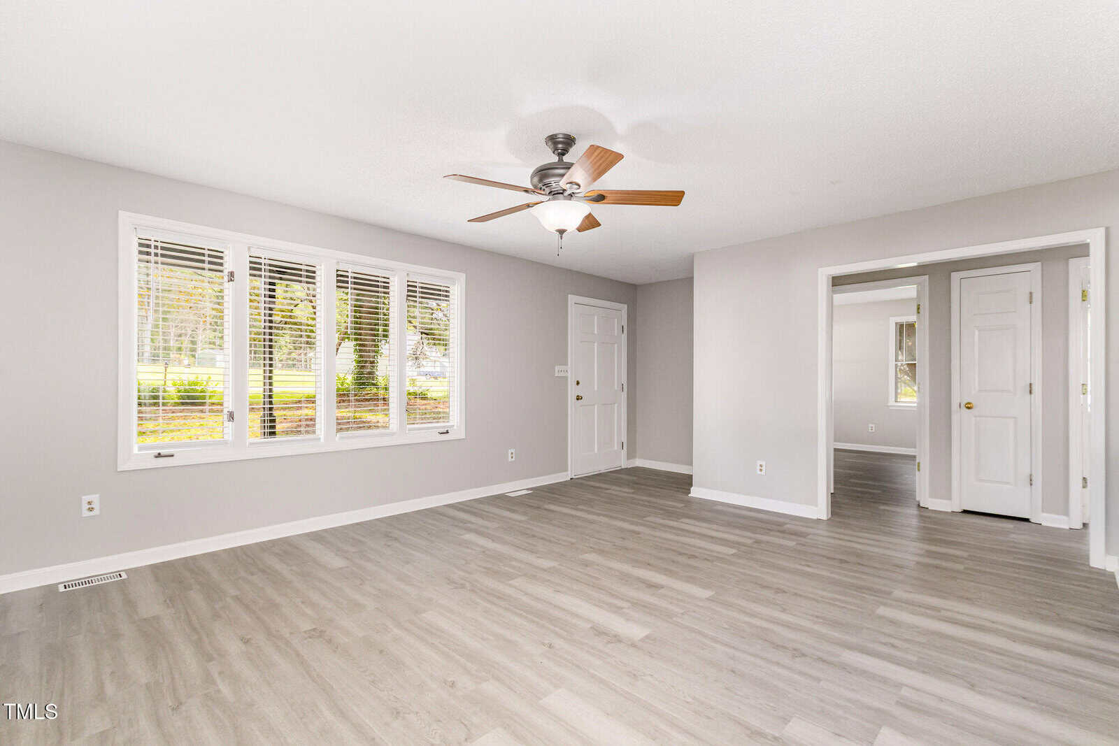 A bright, empty living room with light gray walls, wood-look flooring, a ceiling fan, large front window, and open doorways leading to other rooms. Natural light fills the space.