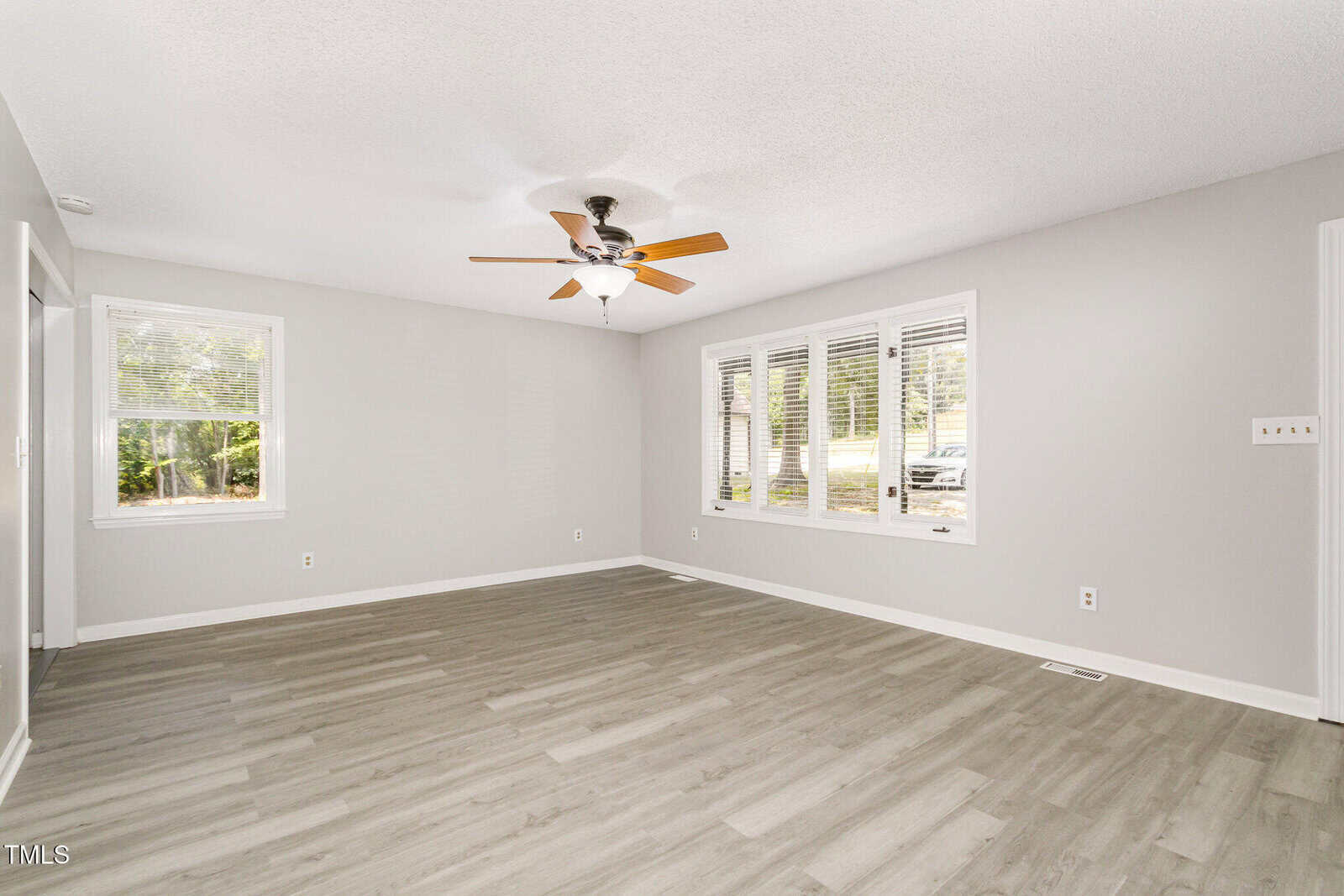 Spacious, empty living room with light gray walls, light wood flooring, a ceiling fan with wooden blades, and large windows letting in natural light.