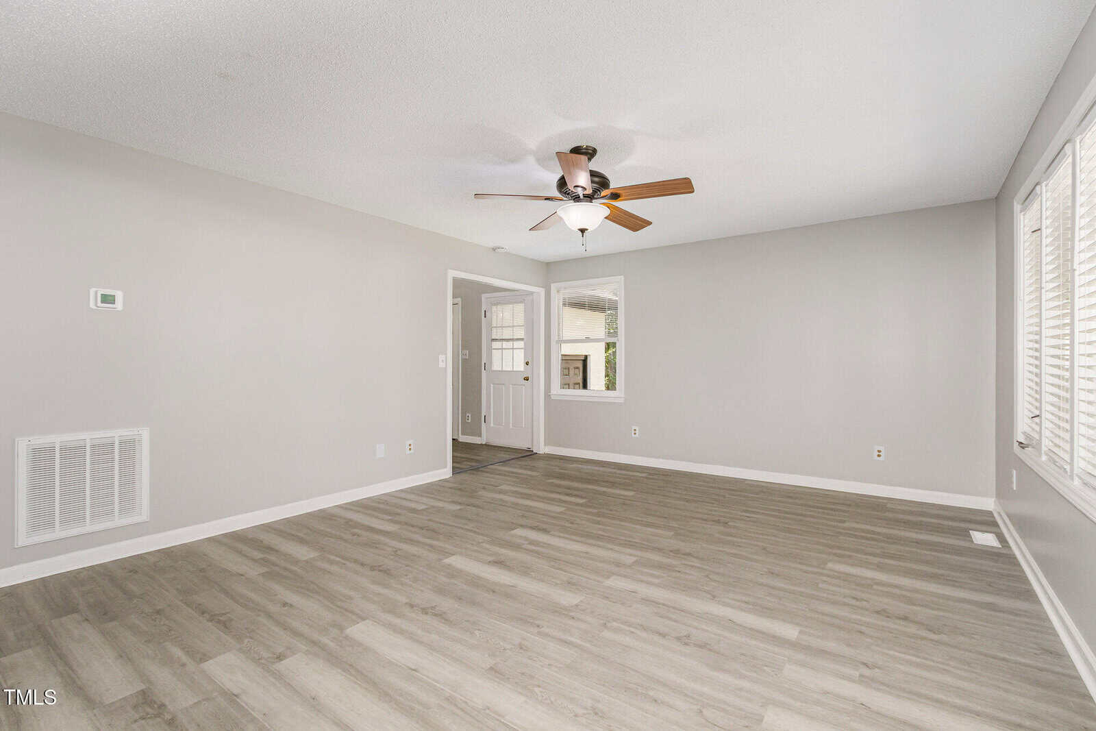 A bright, empty living room with light gray walls, wood-style flooring, a ceiling fan with light, and large windows. A white door and small entryway are visible in the background.