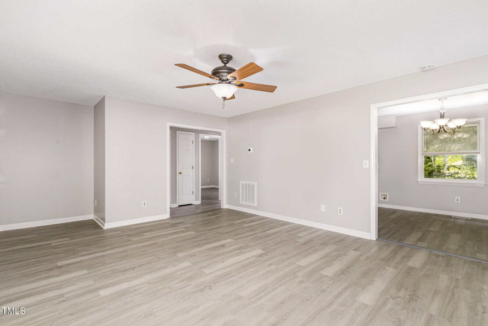 A bright, empty living room with light wood floors, beige walls, a ceiling fan with lights, and an open view into a dining area with a window and chandelier.