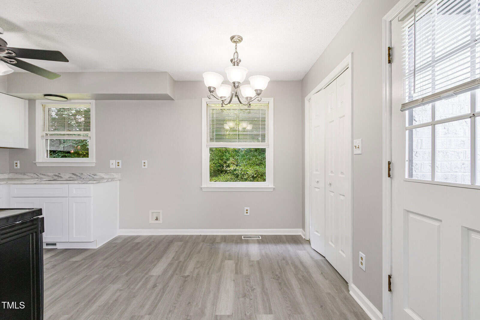 Bright kitchen with light gray walls and floor, white cabinets, and a marble countertop. A window and glass door let in natural light. A chandelier hangs over an empty dining area.