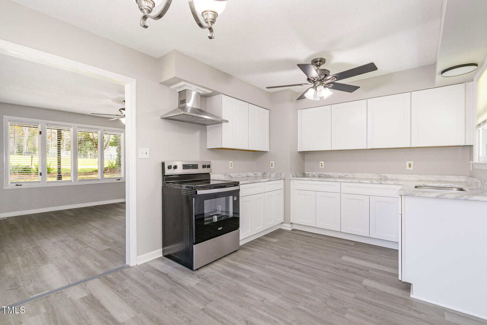 Modern kitchen with white cabinets, stainless steel range, gray countertops, and light wood flooring. Ceiling fans and large windows provide natural light. Adjacent room visible through open doorway.