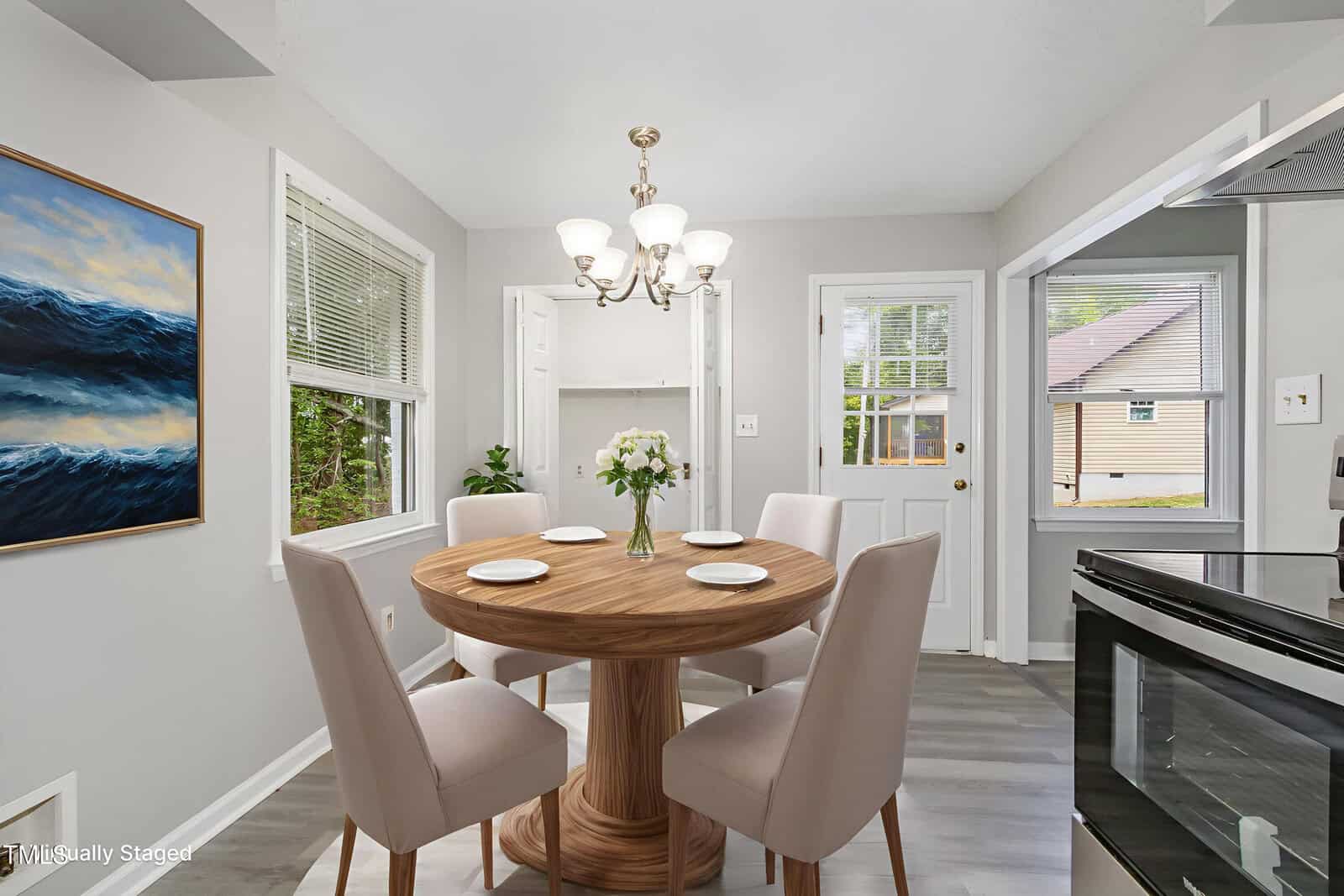 A bright dining area with a round wooden table set for four, beige upholstered chairs, a vase of flowers, large windows, a chandelier, and a view of a house through the back door.
