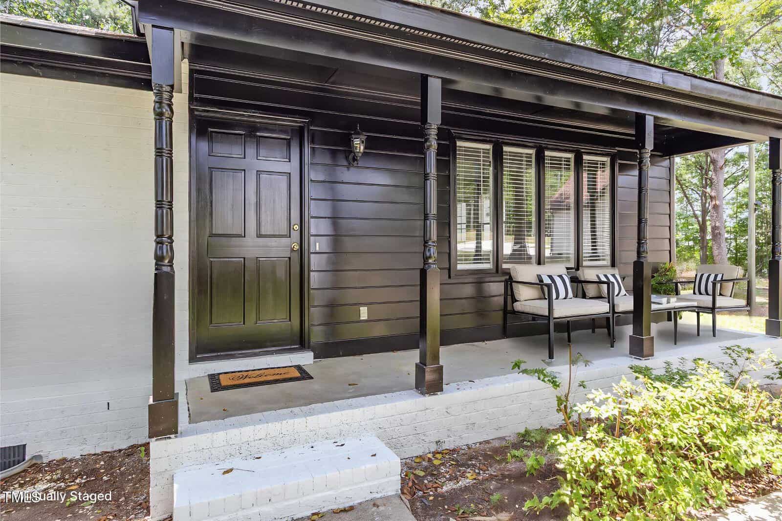 A front porch with dark-painted wood siding, matching front door, two black-and-white cushioned benches, white brick steps, and railing posts; surrounded by greenery and partially shaded by trees.