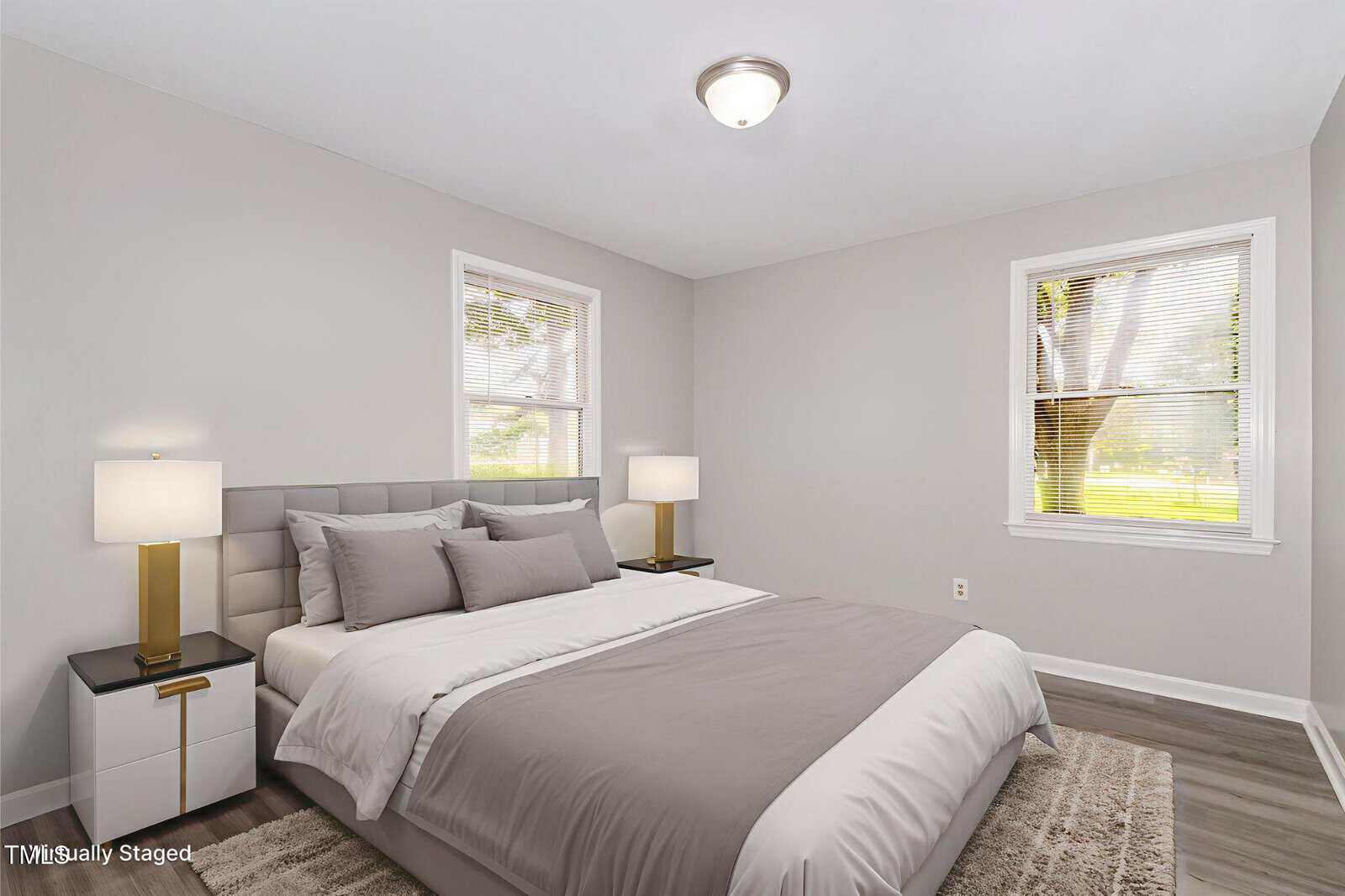 A bright, modern bedroom with a double bed, gray headboard, gray and white bedding, two nightstands with lamps, light gray walls, wood floors, and two windows letting in natural light.