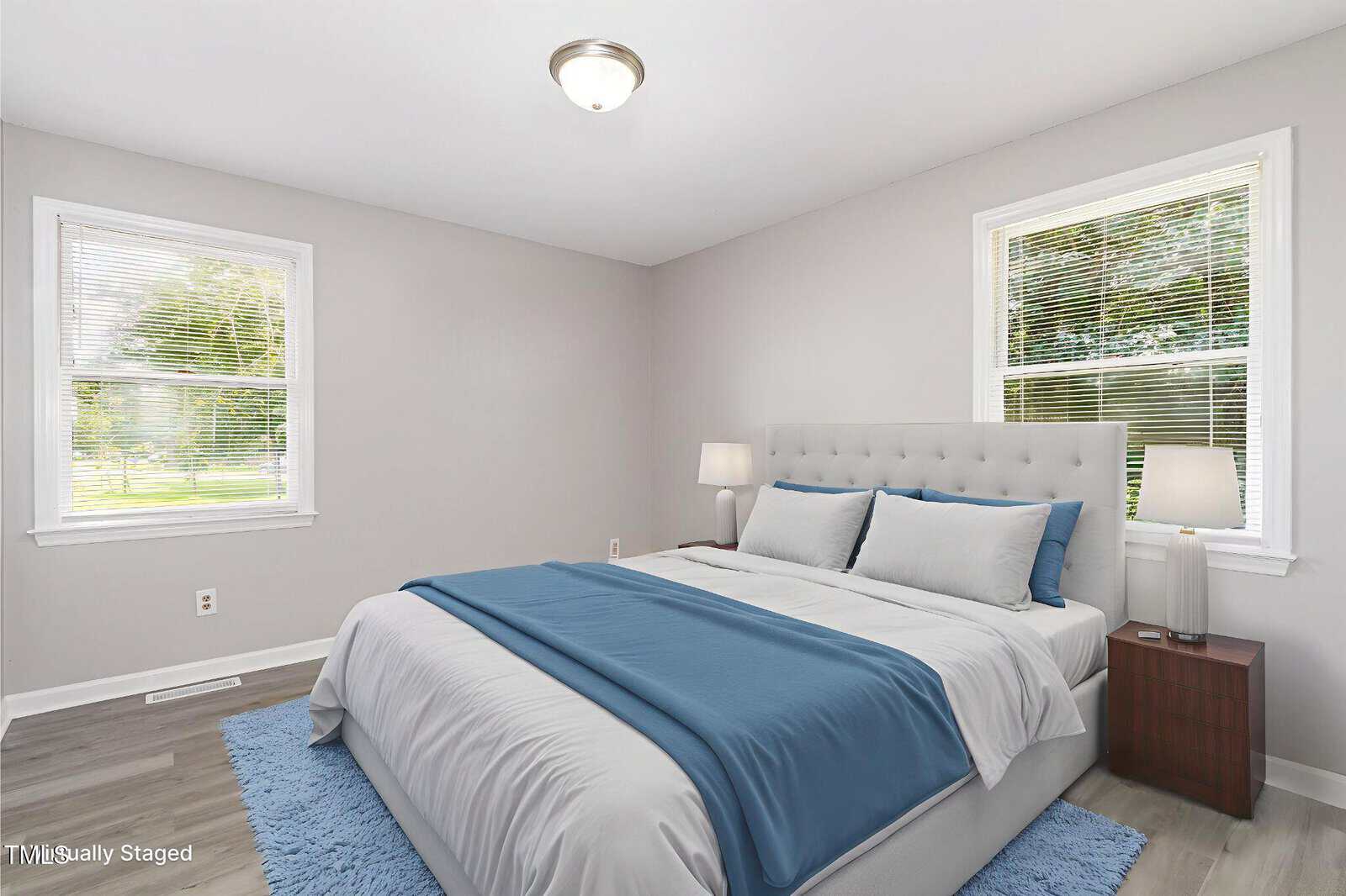 A bright bedroom with light gray walls, two windows with blinds, a large bed with white and blue bedding, two bedside tables with lamps, and a blue rug on the floor.