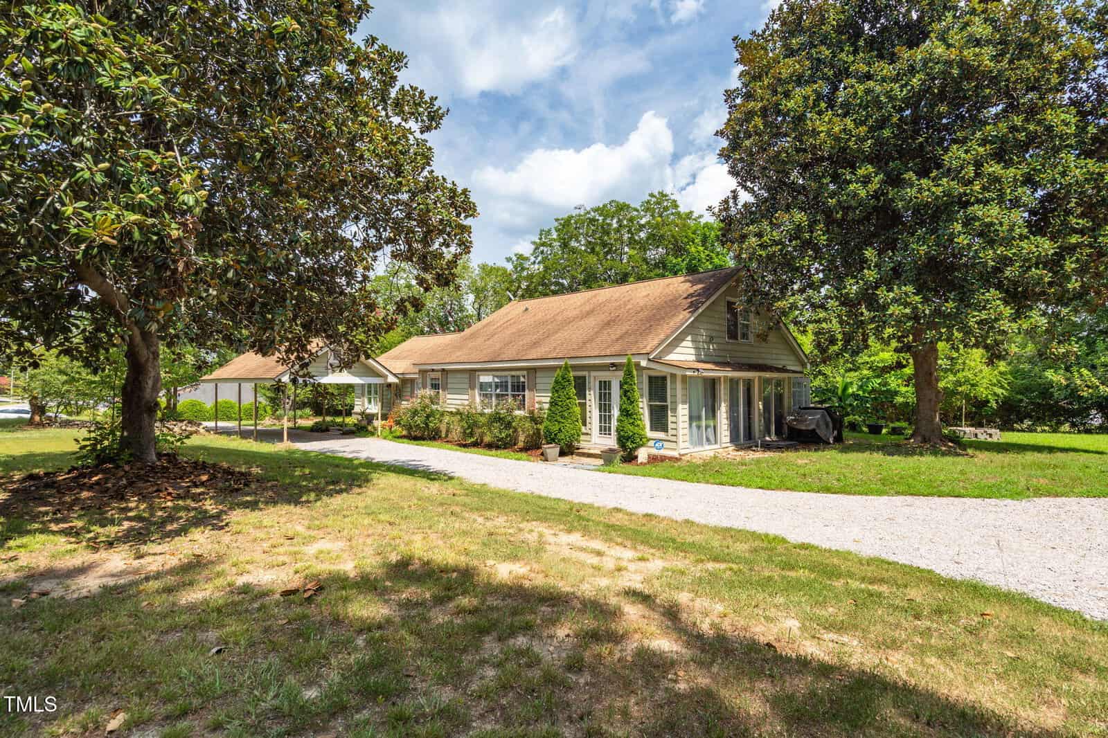 A tan, single-story house with a sloped roof and several large windows sits beside a gravel driveway, surrounded by green trees and a grassy yard on a sunny day.