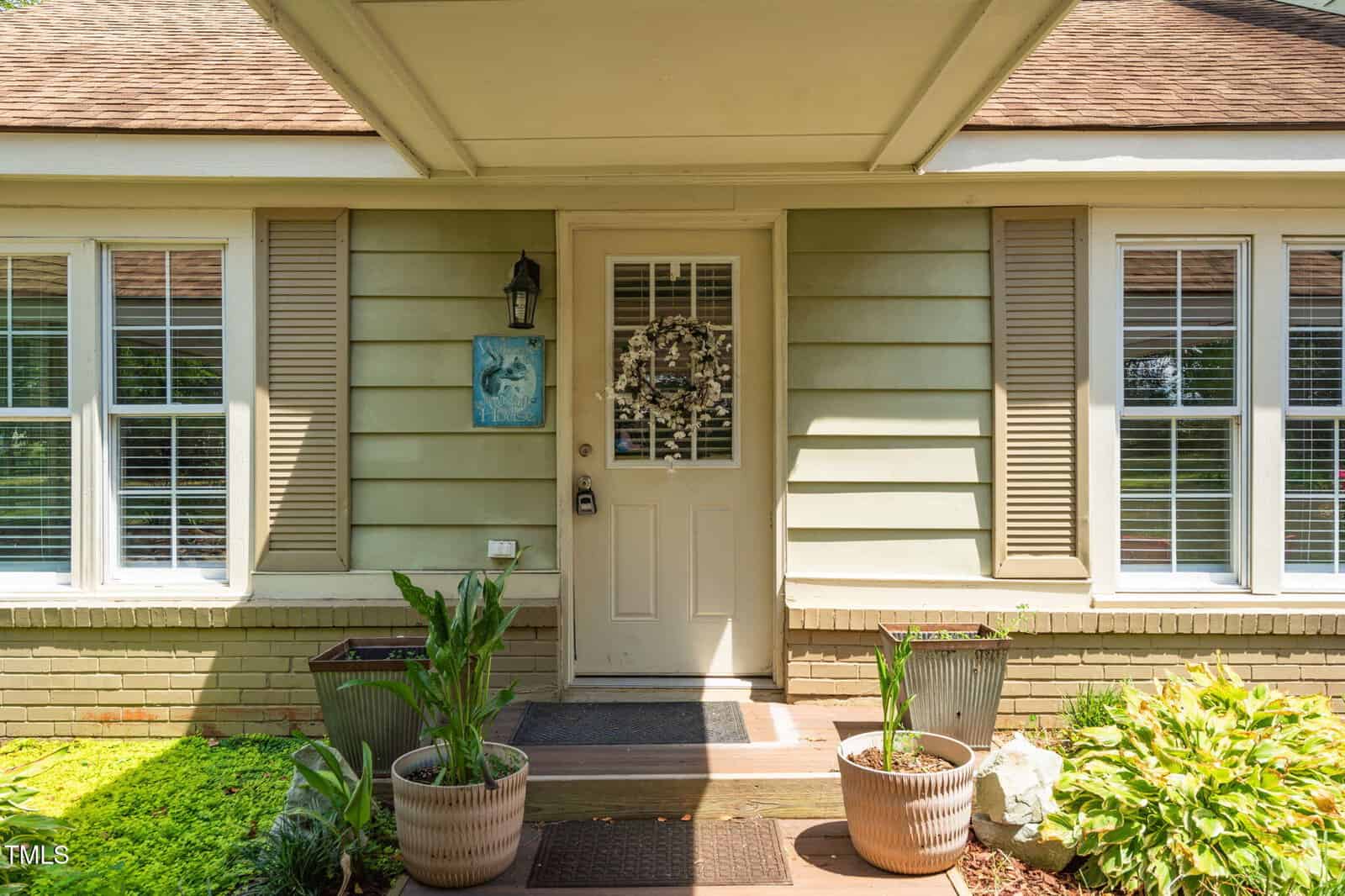 A beige house front features a door with a wreath, two large potted plants on either side of the entry, and windows with closed blinds flanking the door. The house has light green siding and tan shutters.