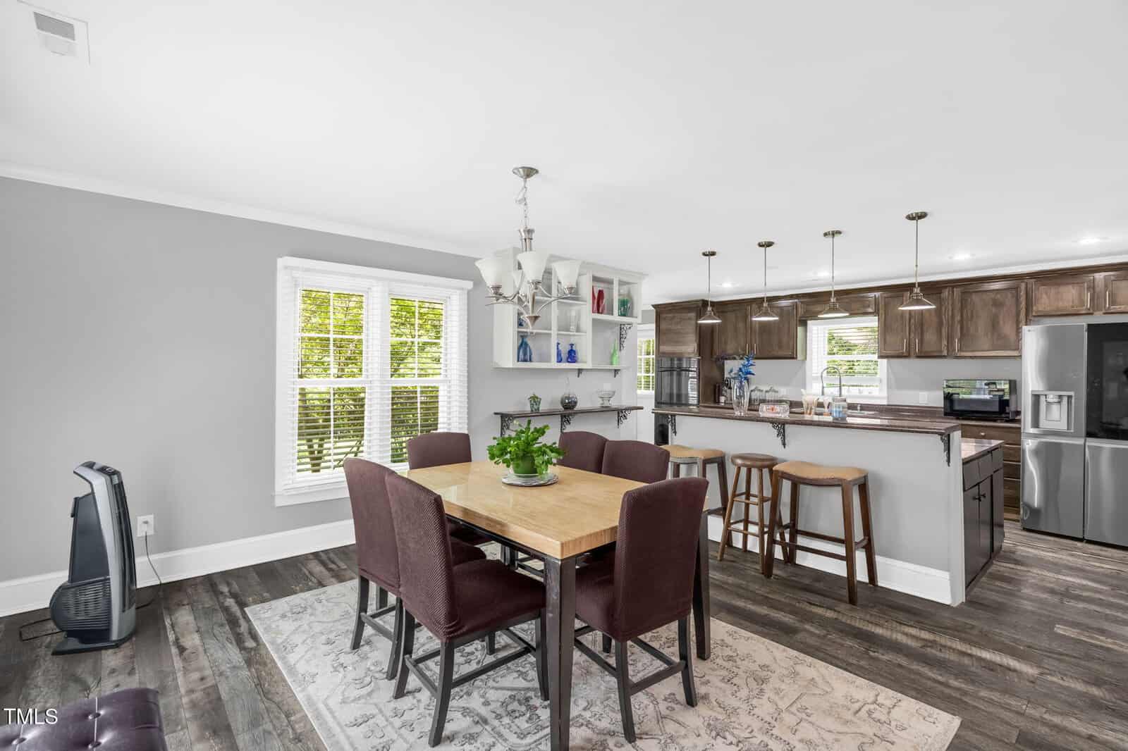 A dining area with a wooden table and six chairs sits next to a kitchen with bar stools, pendant lights, stainless steel appliances, and brown cabinets. A window with blinds lets in natural light.