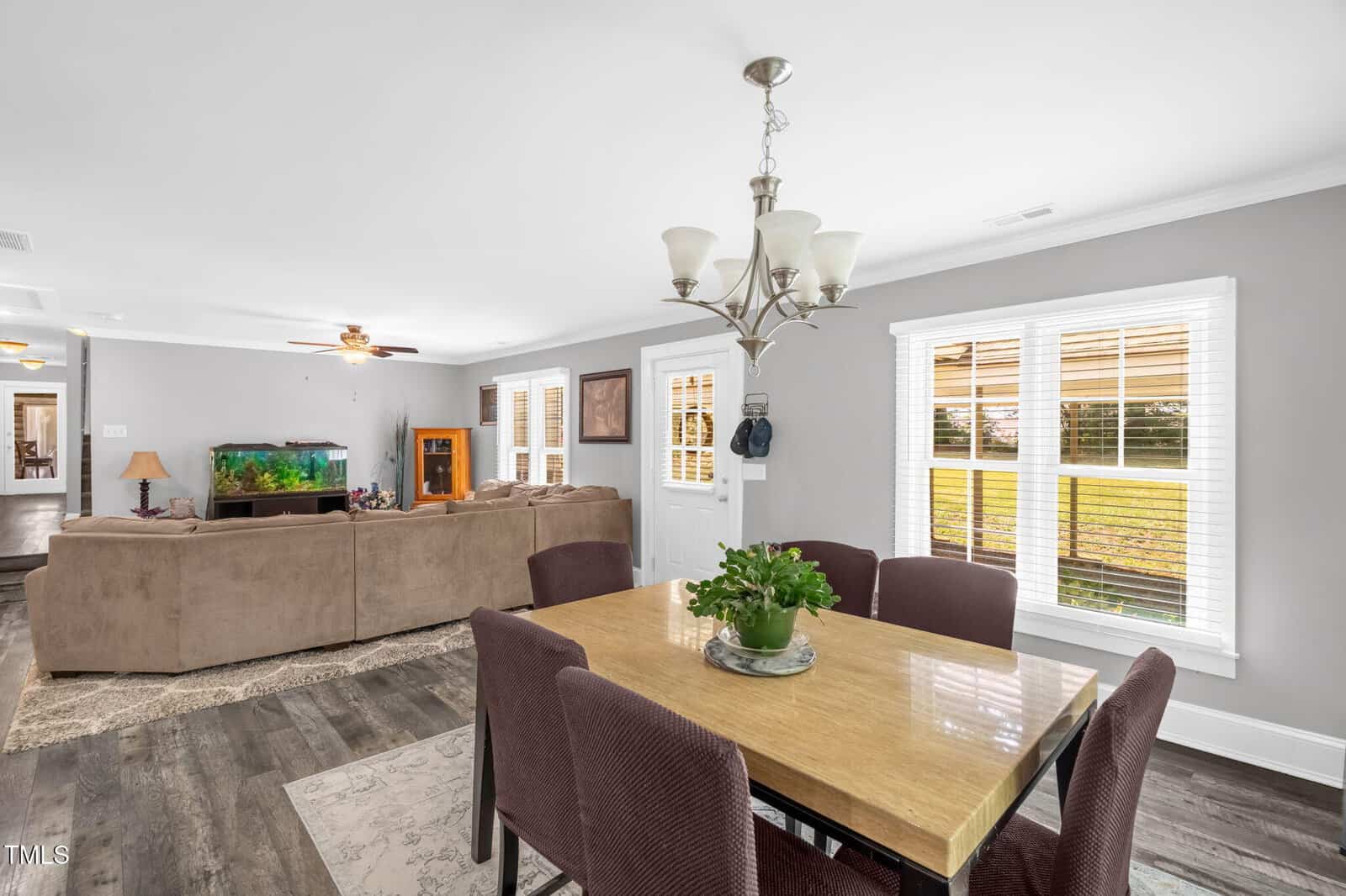 Open living and dining area with a beige sectional sofa, fish tank, and a wooden dining table with six purple chairs. Large windows let in natural light. Walls are light gray; flooring is dark wood. A light fixture hangs above the table.