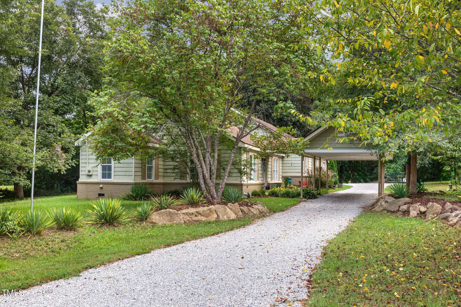 A light green house with a gabled roof and attached carport sits behind leafy trees and a white gravel driveway, surrounded by green grass, shrubs, and rocks on a summer day.