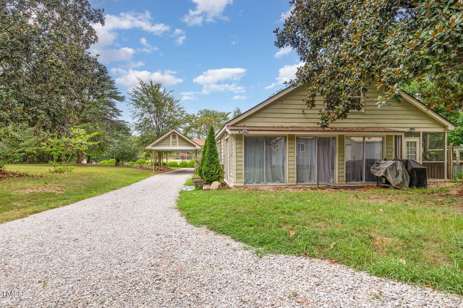 Single-story house with green siding, large windows, and a gravel driveway. There is a barbecue grill on the patio, green grass, trees, and a smaller outbuilding in the background under a blue sky with scattered clouds.