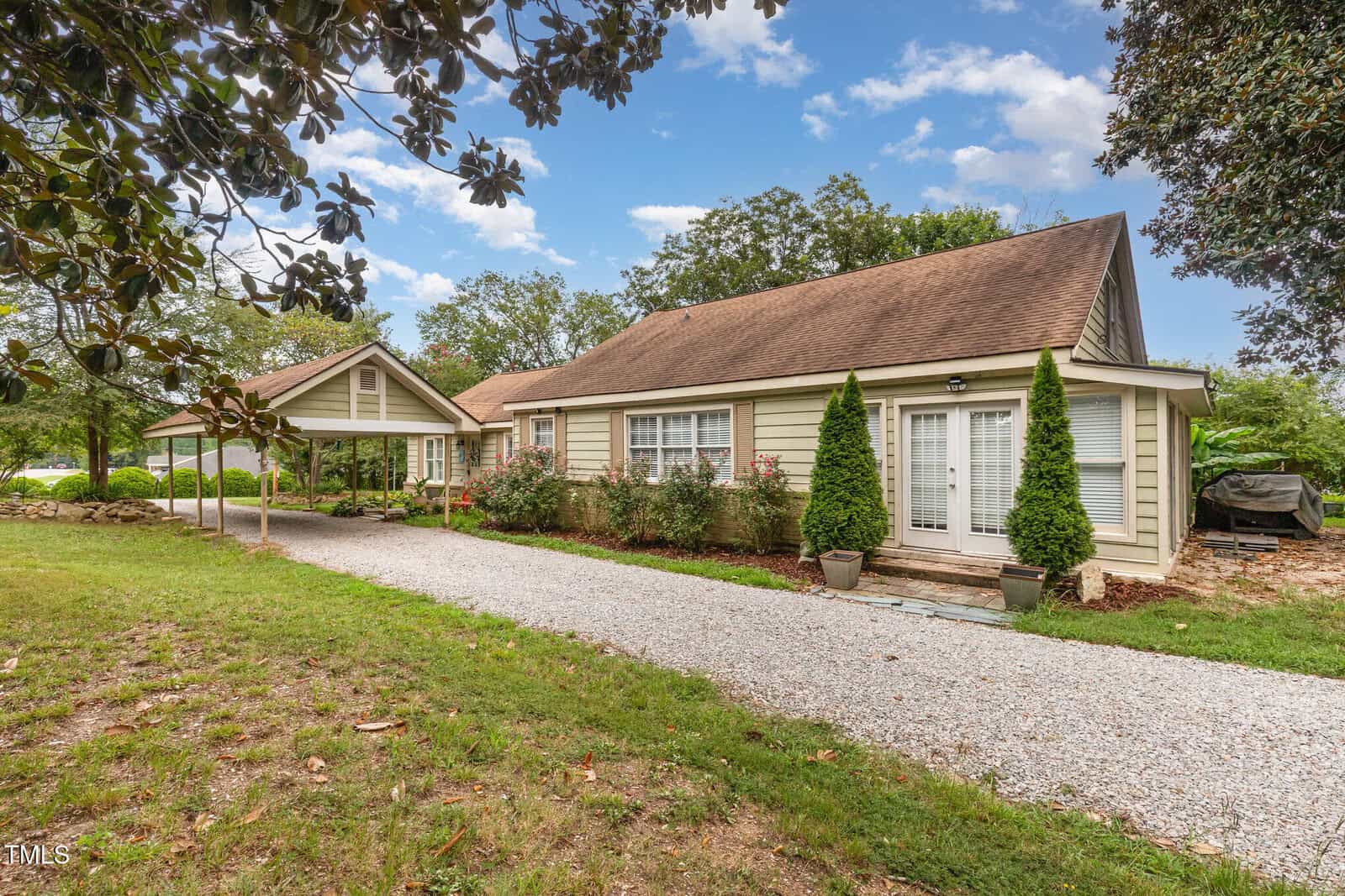 Single-story beige house with white trim, large windows, and a brown roof. There is a gravel driveway, two potted shrubs by French doors, and a covered carport surrounded by trees and greenery.