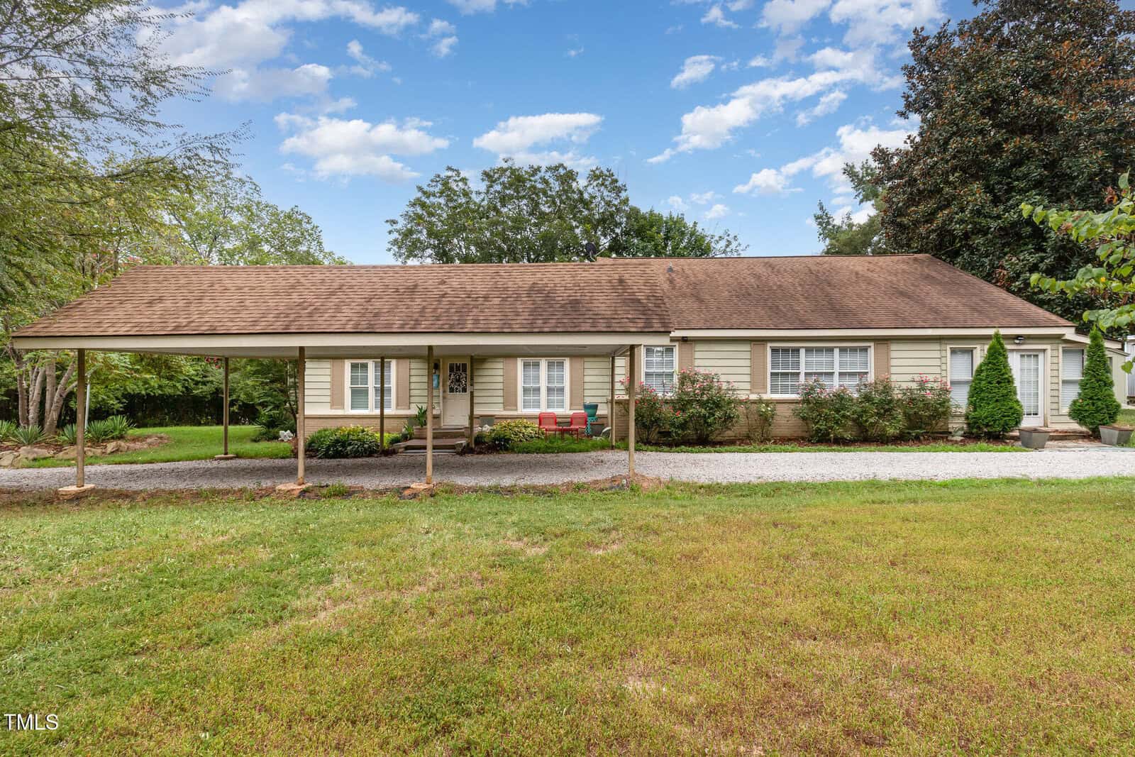 Single-story beige house with brown shingle roof, attached carport on the left, front porch with chairs, gravel driveway, green lawn, trees, and blue sky with scattered clouds in the background.