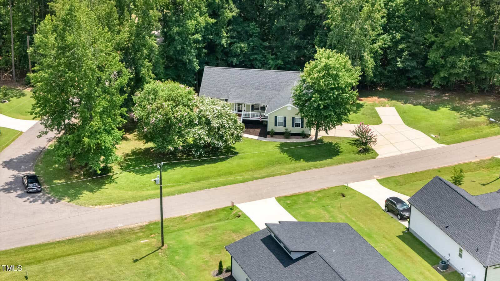 A single-story house with a front porch sits on a large grassy lot with a curved driveway, surrounded by trees and other homes, bordered by a street.