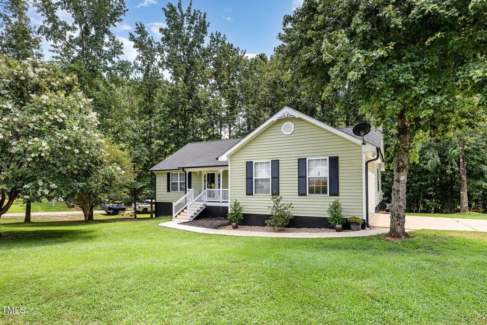 Single-story house with light yellow siding, black shutters, a covered front porch, and driveway. The lawn is well-maintained and surrounded by trees and shrubs under a partly cloudy sky.
