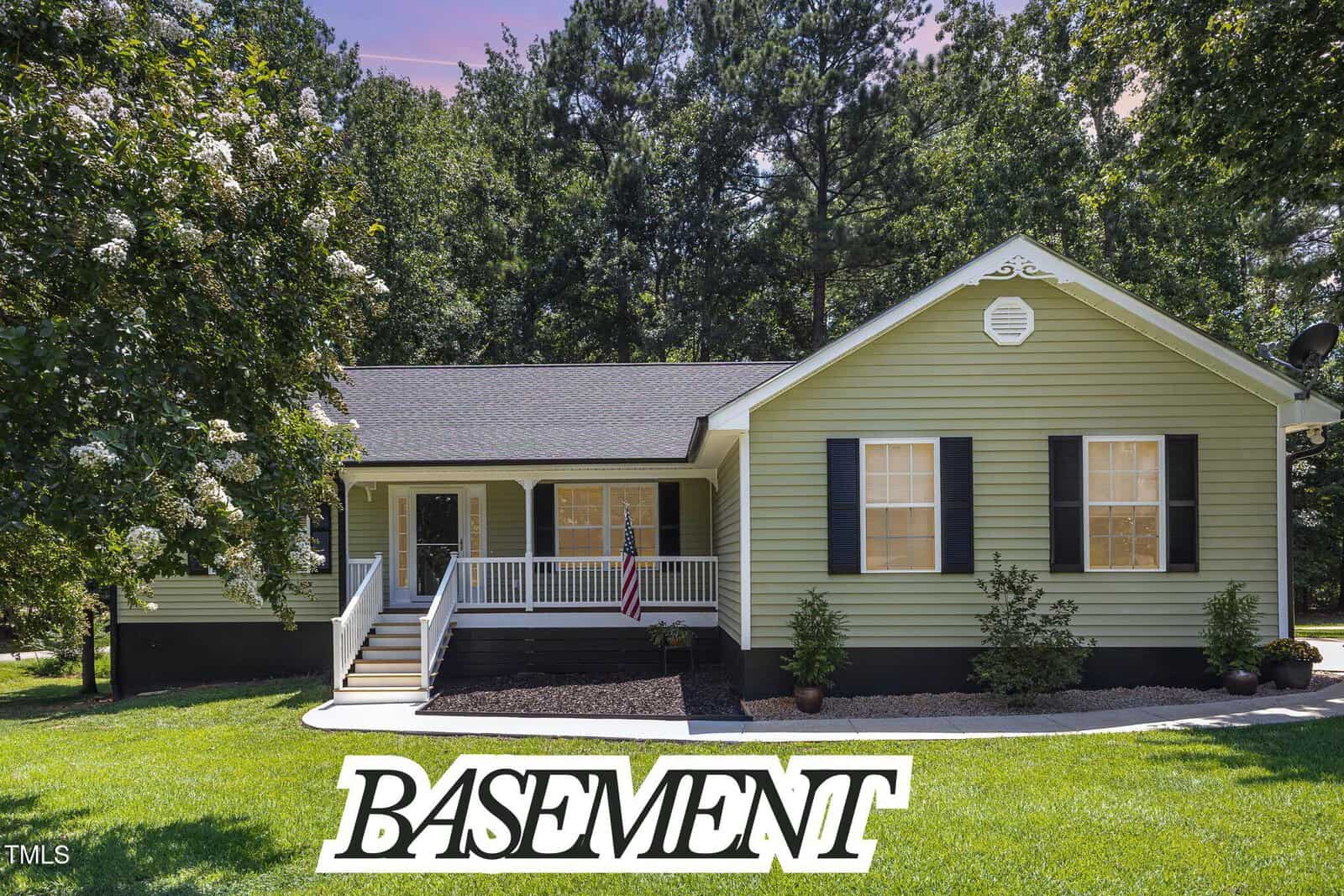 Single-story green house with white trim, black shutters, and front porch, set on a grassy yard with trees in the background at 144 Ottawa Drive, Louisburg. The word “BASEMENT” is written in large white text at the bottom of the image.