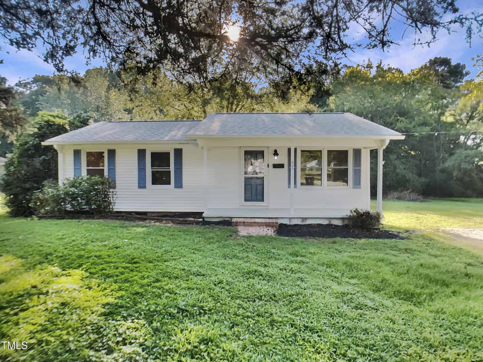 Charming and welcoming, this single-story white house at 605 Mitchell Avenue in Franklinton features blue shutters, a dark blue front door, and a covered porch. The home is nestled among lush green grass, bushes, and trees, with sunlight filtering through the branches overhead.