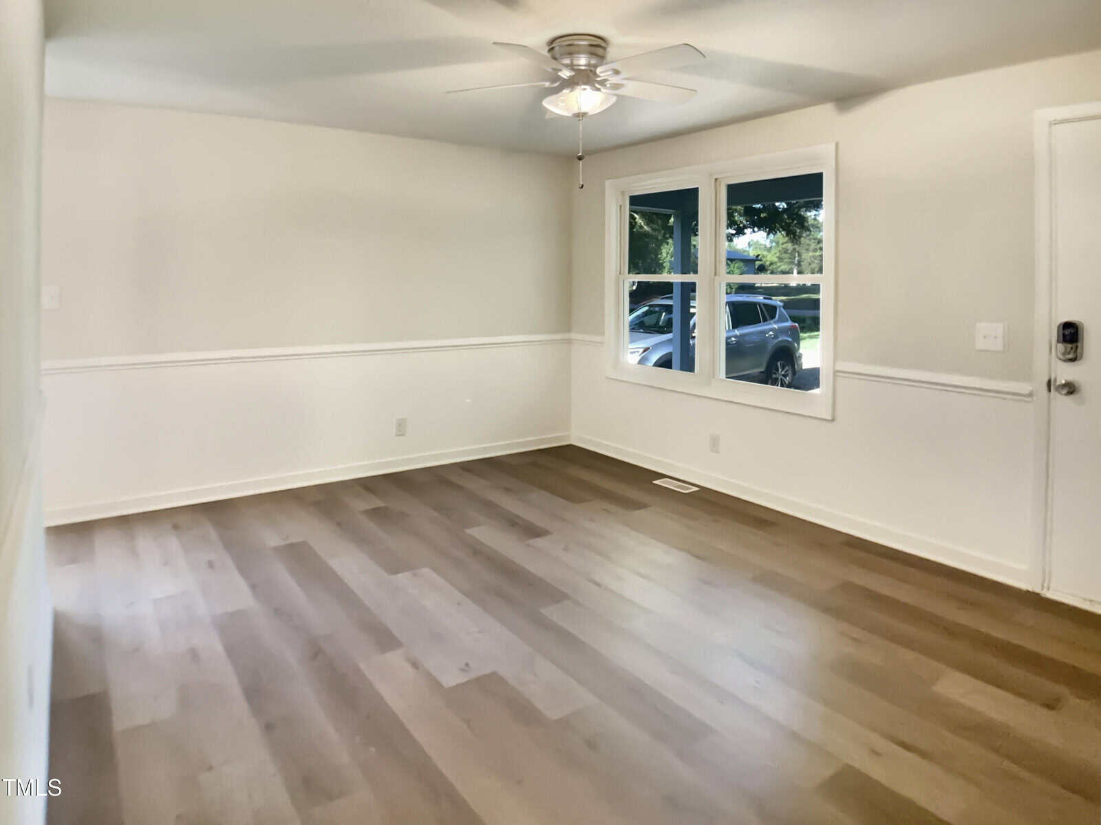 Empty room with light-colored walls, wood flooring, a white ceiling fan with a light, two windows showing parked cars and greenery outside, and a white door to the right of the windows.