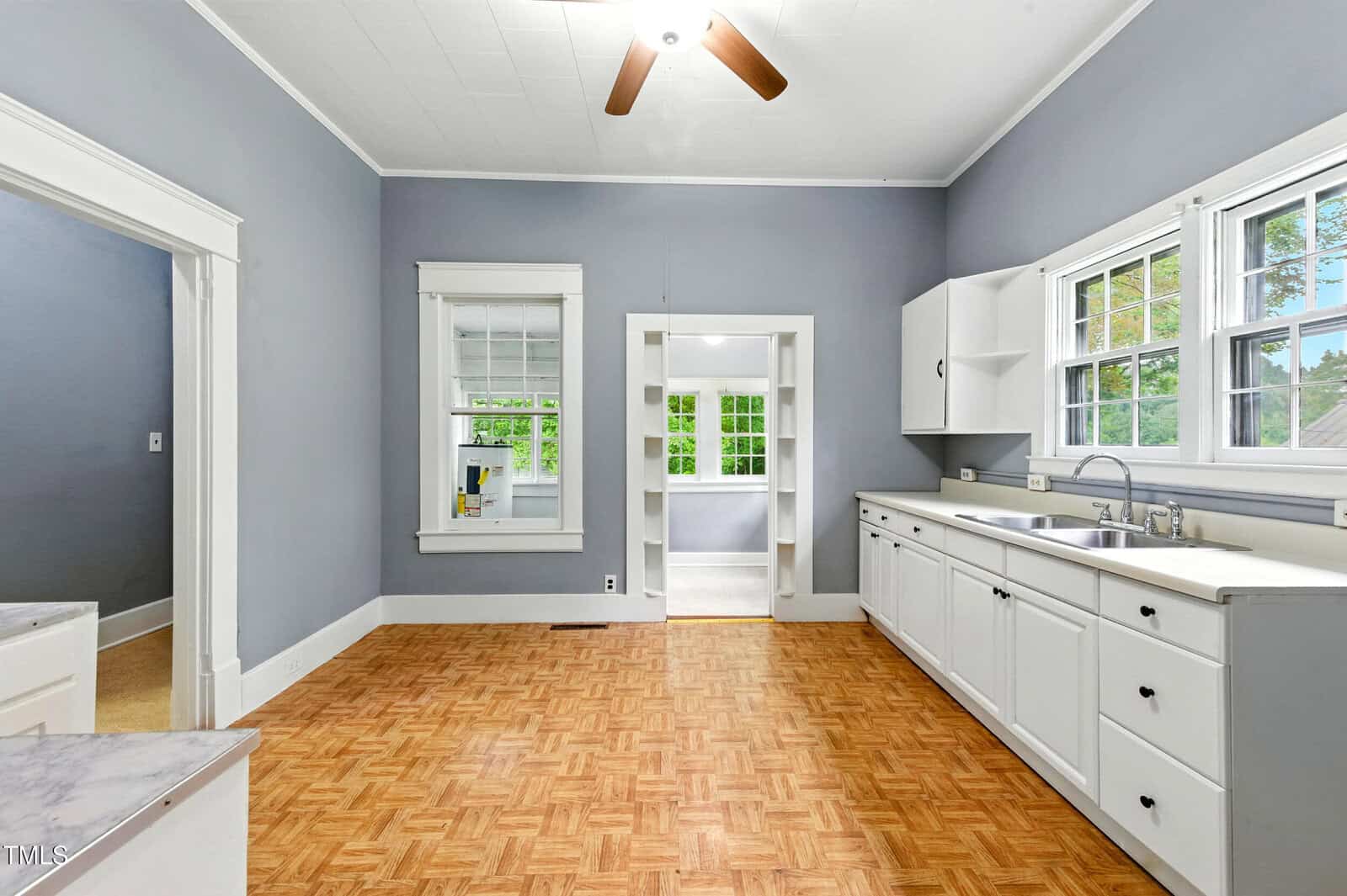 A spacious kitchen with gray walls, white cabinets, parquet wood flooring, a ceiling fan, and multiple windows letting in natural light. An adjacent room is visible through an open doorway.