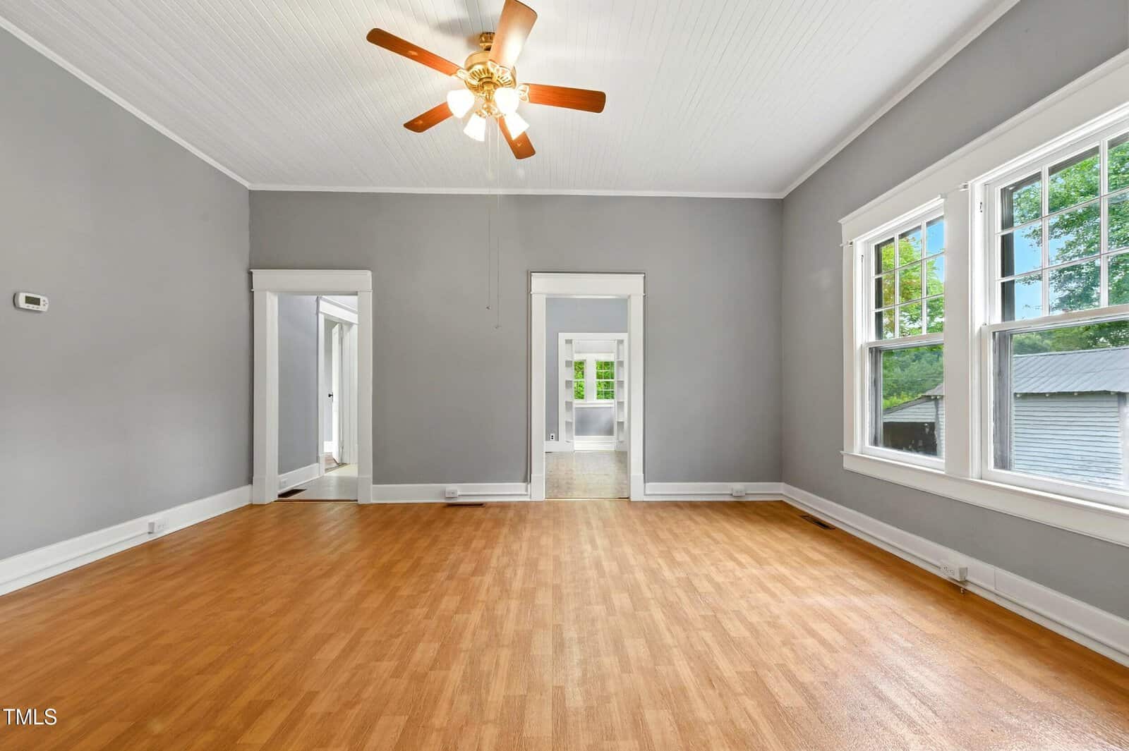 A spacious, empty room with light wood flooring, gray walls, white trim, large windows on the right, a ceiling fan with lights, and doorways leading to adjacent rooms.