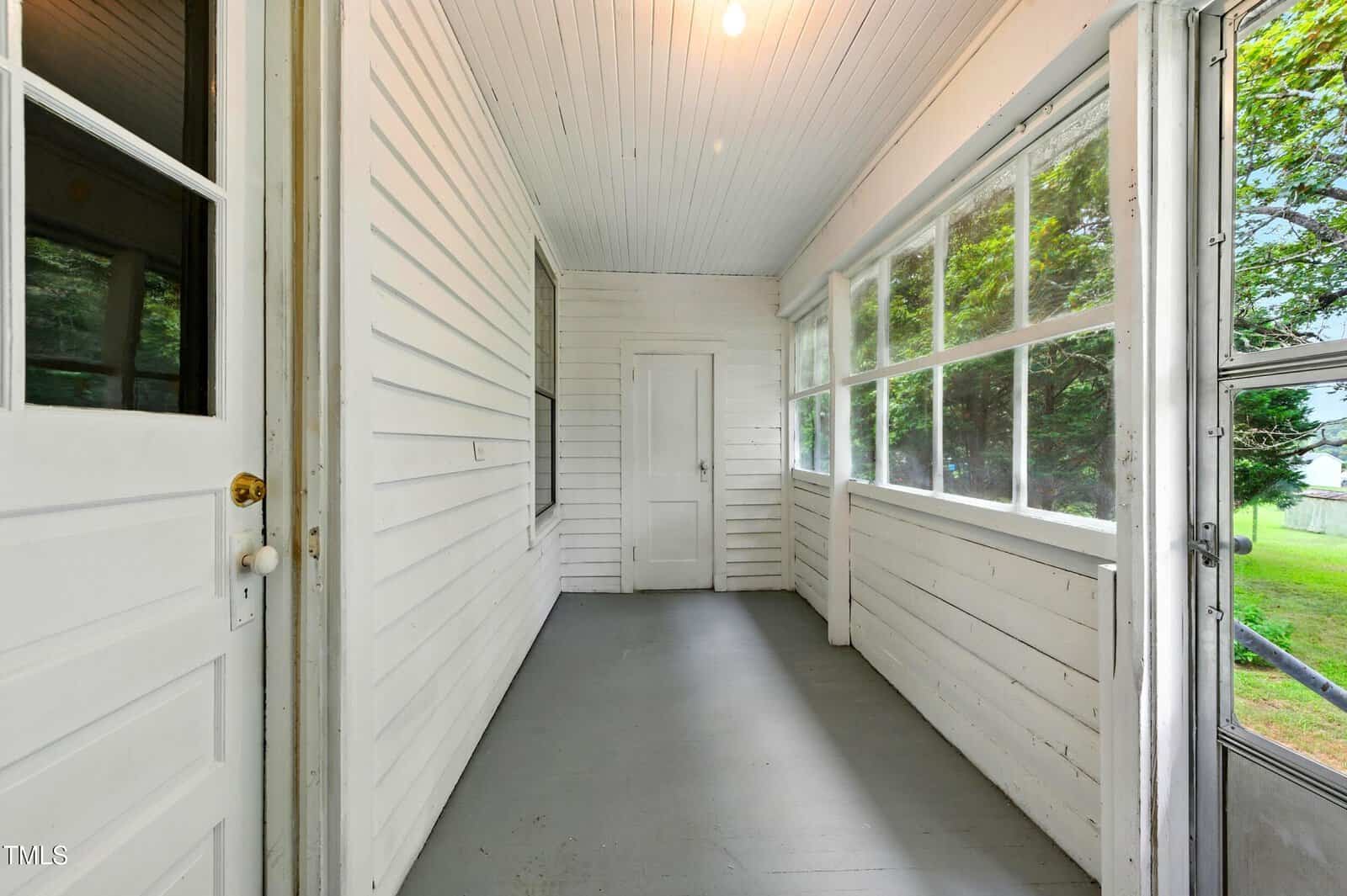 A white, enclosed porch with large windows, a gray floor, and a door at the far end, letting in natural light and showing green trees outside.