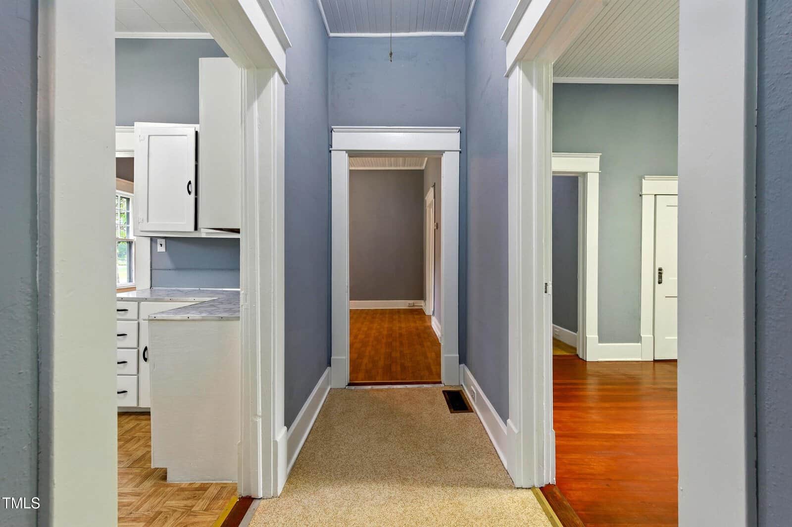 A hallway with carpet flooring leads to rooms with hardwood floors and white trim. The left shows a kitchen with white cabinets; the right shows another room. Walls are painted a muted blue-gray color.
