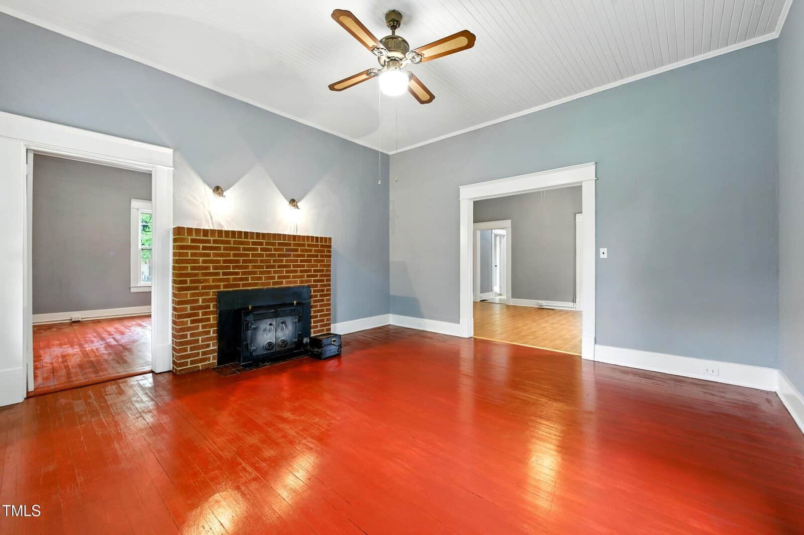 Living room with red-stained wood floors, a brick fireplace with a black insert, gray walls, white trim, and a ceiling fan with lights. Doorways lead to adjacent rooms with similar decor and wood floors.