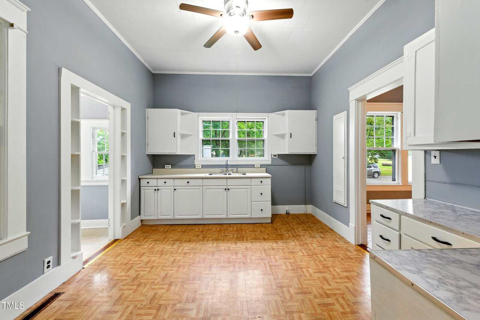 A kitchen with white cabinets, gray walls, wood-patterned flooring, a ceiling fan with lights, and two windows above the sink. An open doorway leads to an adjacent room with tan walls and visible windows.