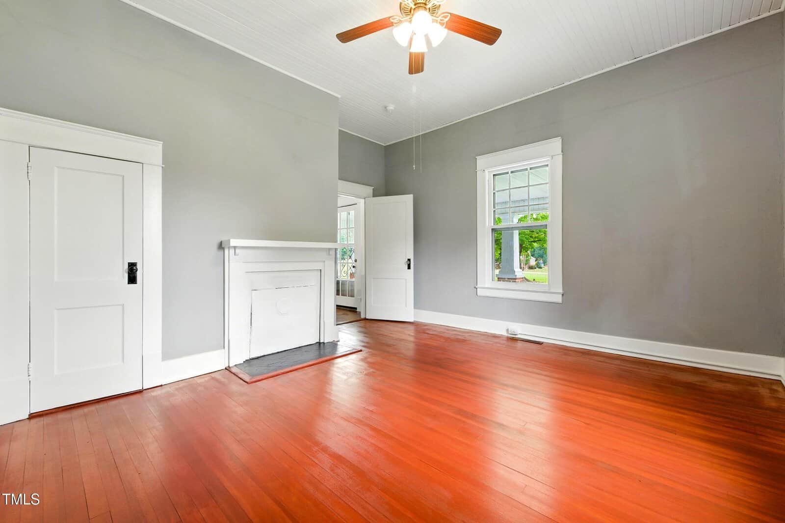 Empty room with reddish wooden floor, light gray walls, white trim, a ceiling fan with lights, a white door, fireplace, and large window showing greenery outside.