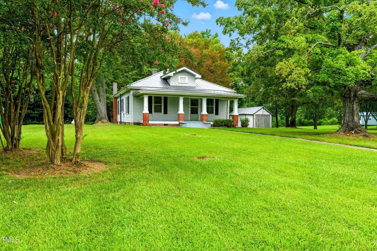 A single-story white house with a gray metal roof, brick porch columns, and a covered front porch sits on a large grassy lawn with mature trees and a small shed in the background. The sky is partly cloudy.