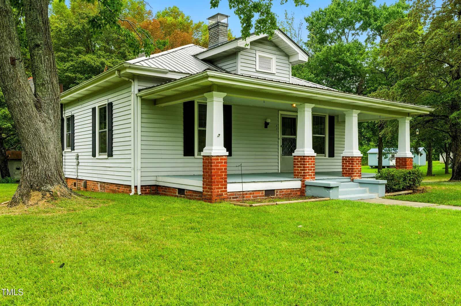 A single-story house with light gray siding, a metal roof, red brick foundation, a covered front porch with white columns, and a lawn with trees in the background.