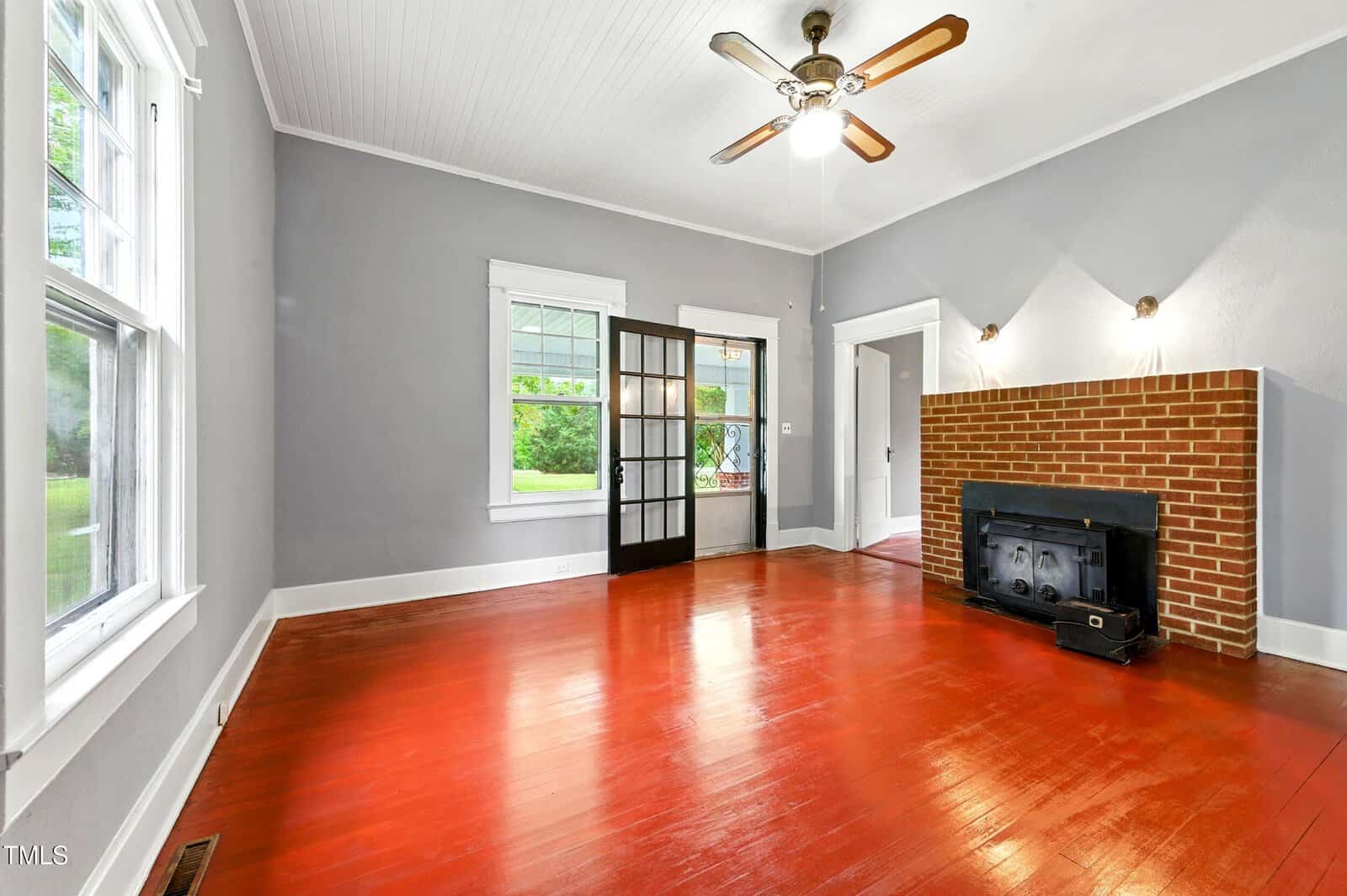 A living room with red hardwood floors, gray walls, a white ceiling, and a ceiling fan. There is a brick fireplace with a wood stove insert and two wall lights above it, and a set of French doors near two large windows.