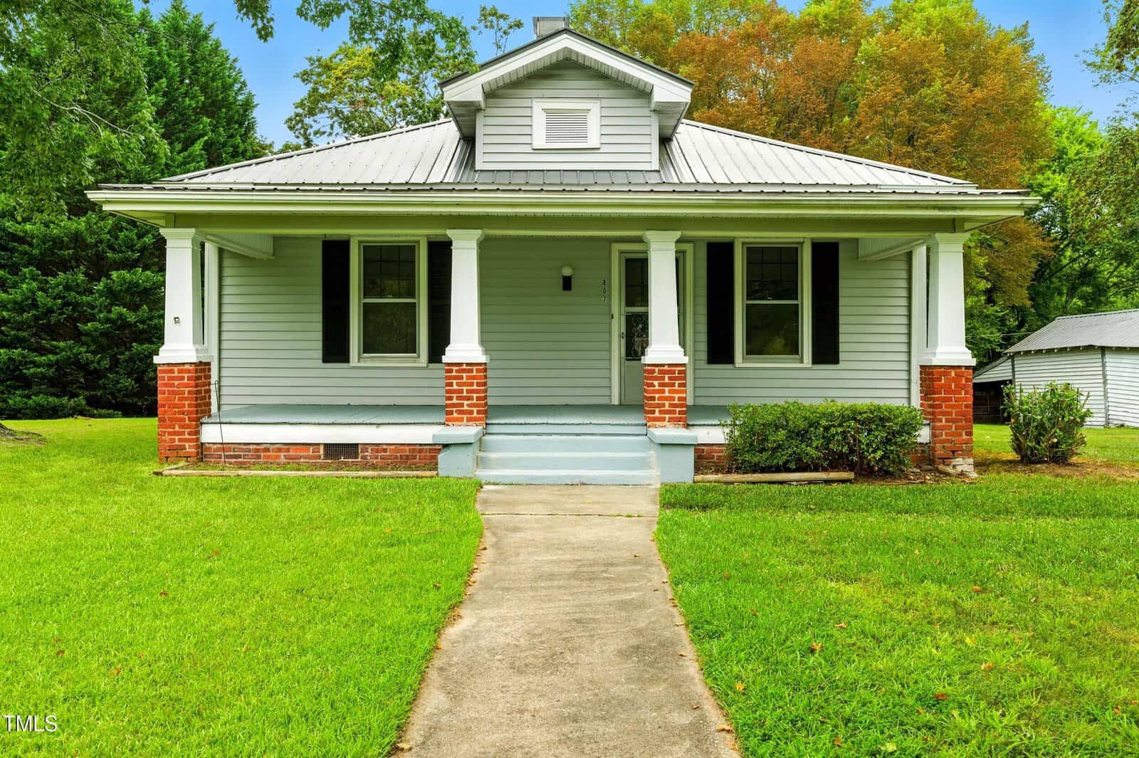 This charming single-story house at 807 N Main Street in Creedmoor features gray siding, white trim, a metal roof, and a welcoming covered front porch with brick columns. The home sits on a lush green lawn with trees and a shed in the background.