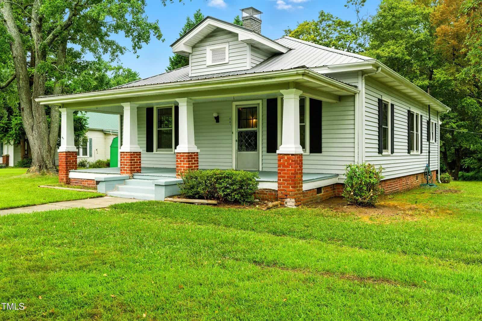 A single-story house with light gray siding, black shutters, a covered front porch supported by white columns with brick bases, and a neatly trimmed lawn surrounded by trees.