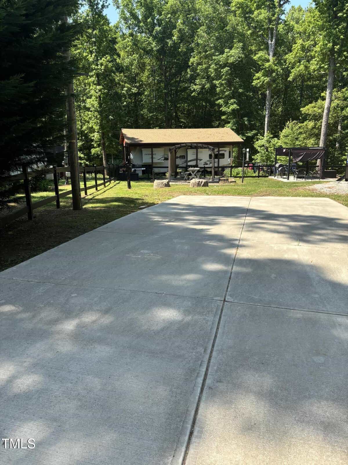 A concrete driveway leads to an RV parked under a small wooden shelter surrounded by trees and greenery on a sunny day.