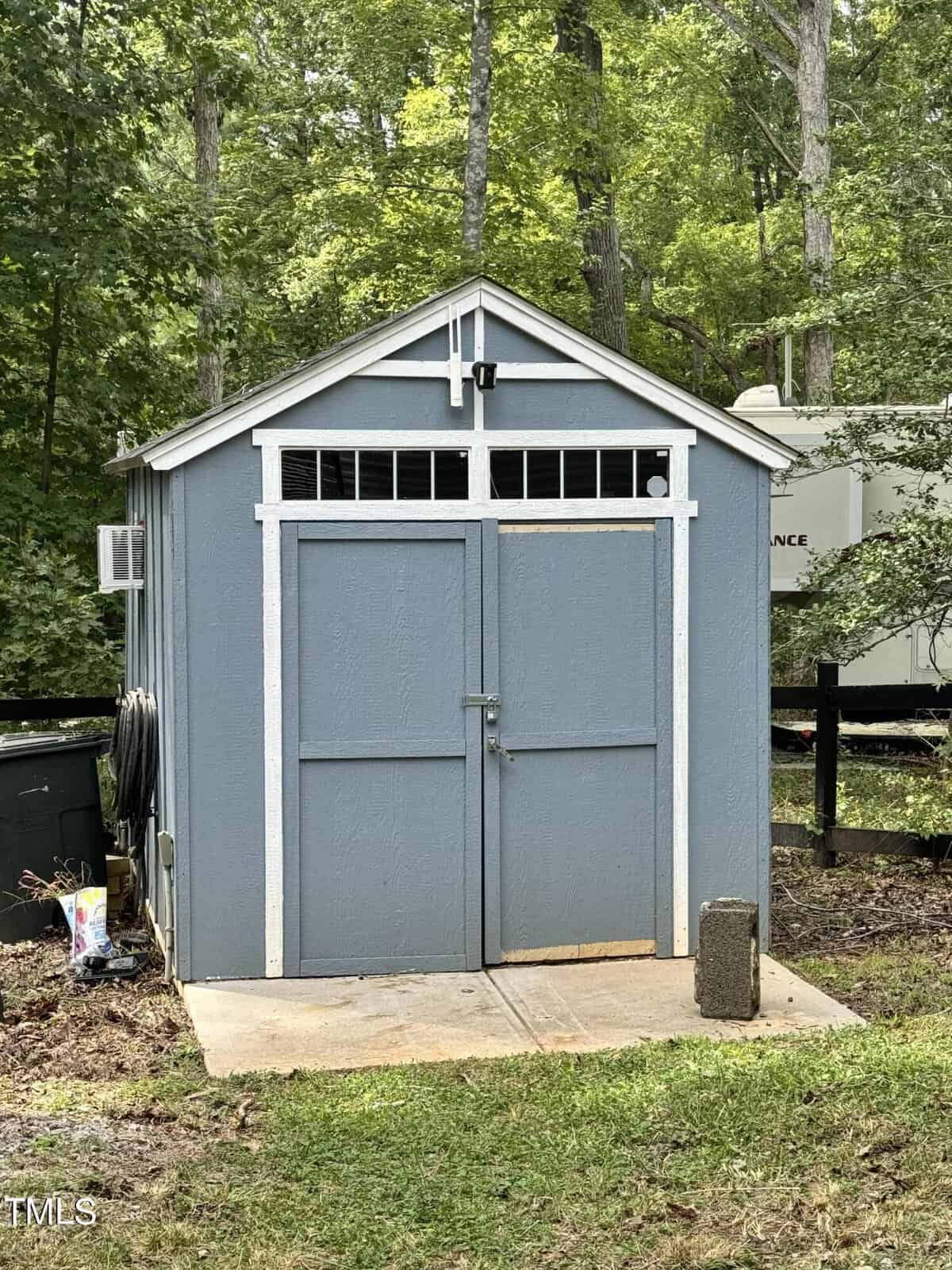 A small blue storage shed with white trim and double doors sits on a concrete pad in a grassy yard, surrounded by trees and outdoor equipment.