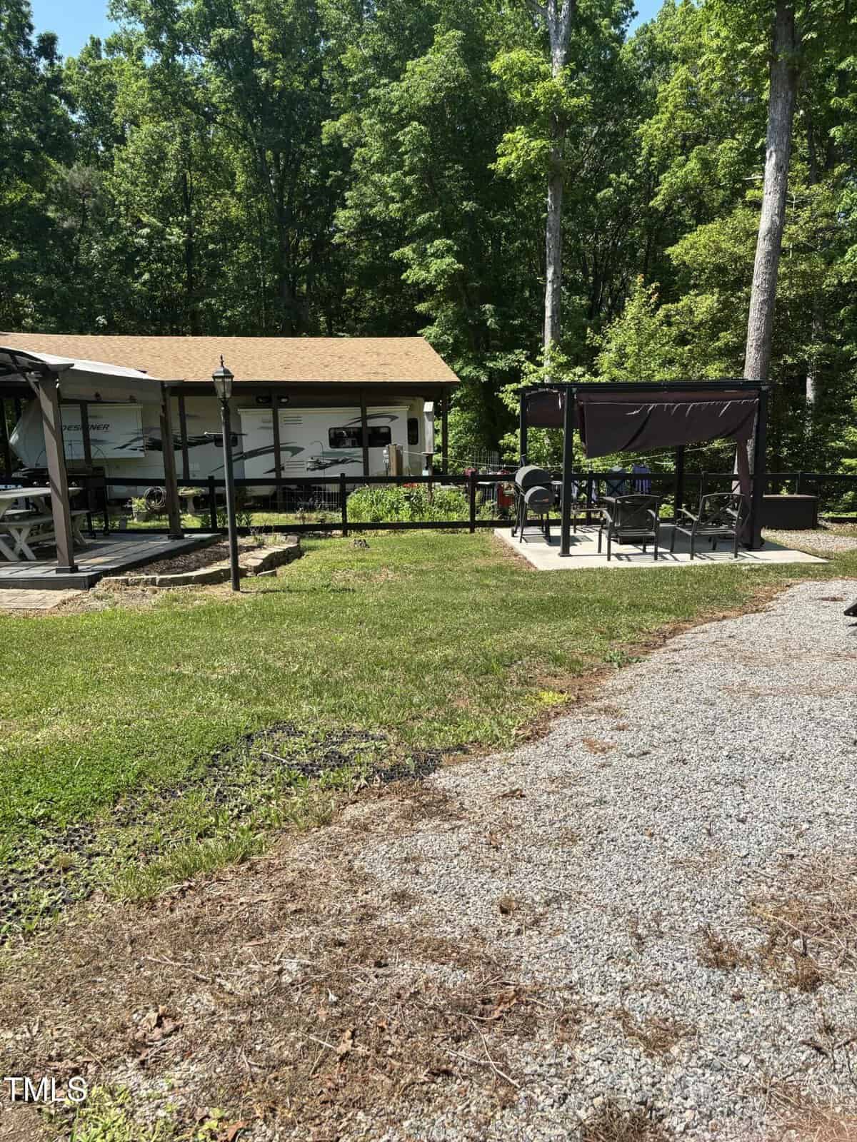 A gravel driveway leads to a grassy yard with a covered picnic area, outdoor seating under a canopy, and an RV parked beside a wooden deck; trees and greenery surround the space.