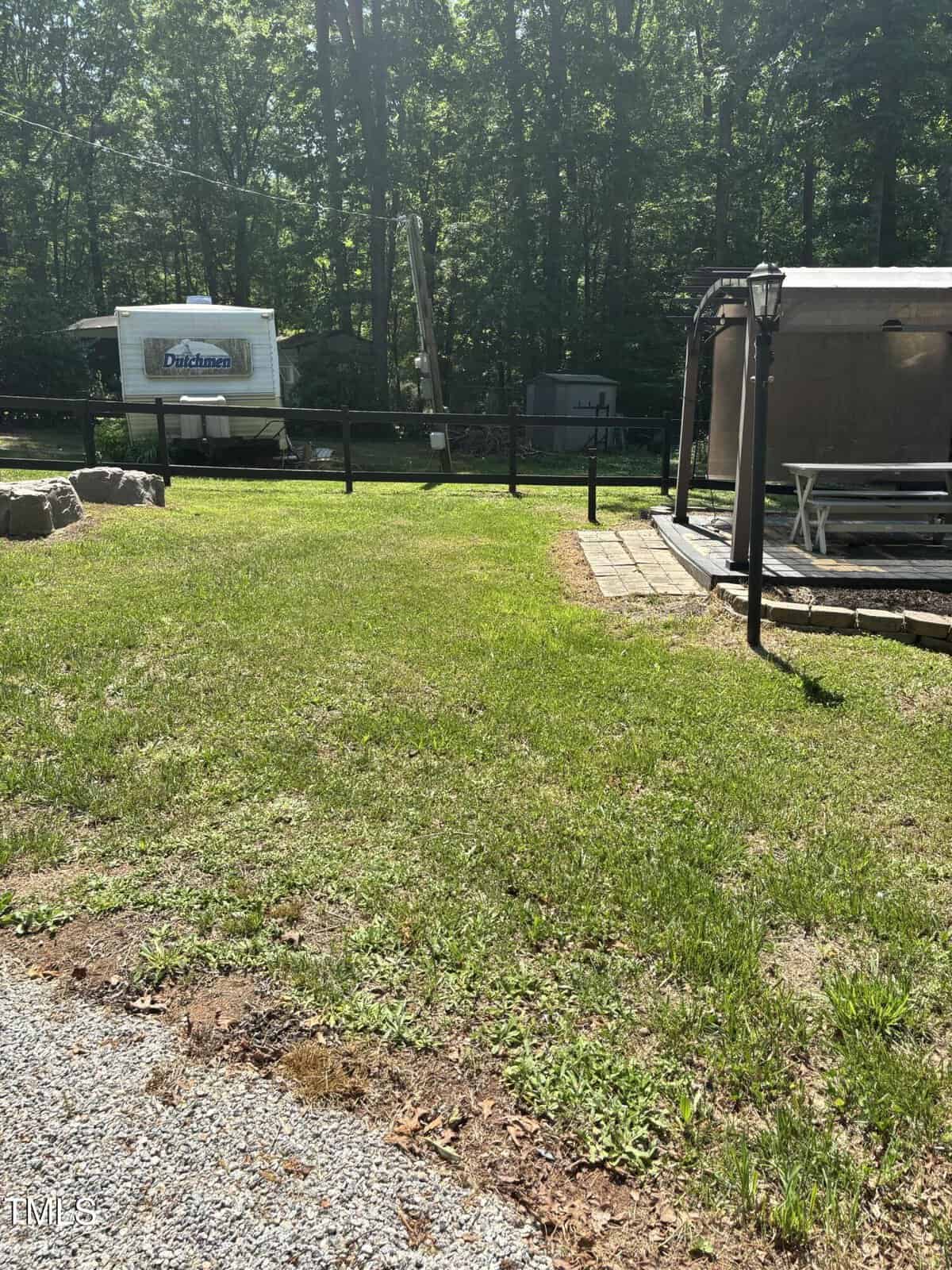 A grassy yard with a picnic table under a canopy, a gravel area in the foreground, and two RVs parked in the background near a wooded area under sunlight.