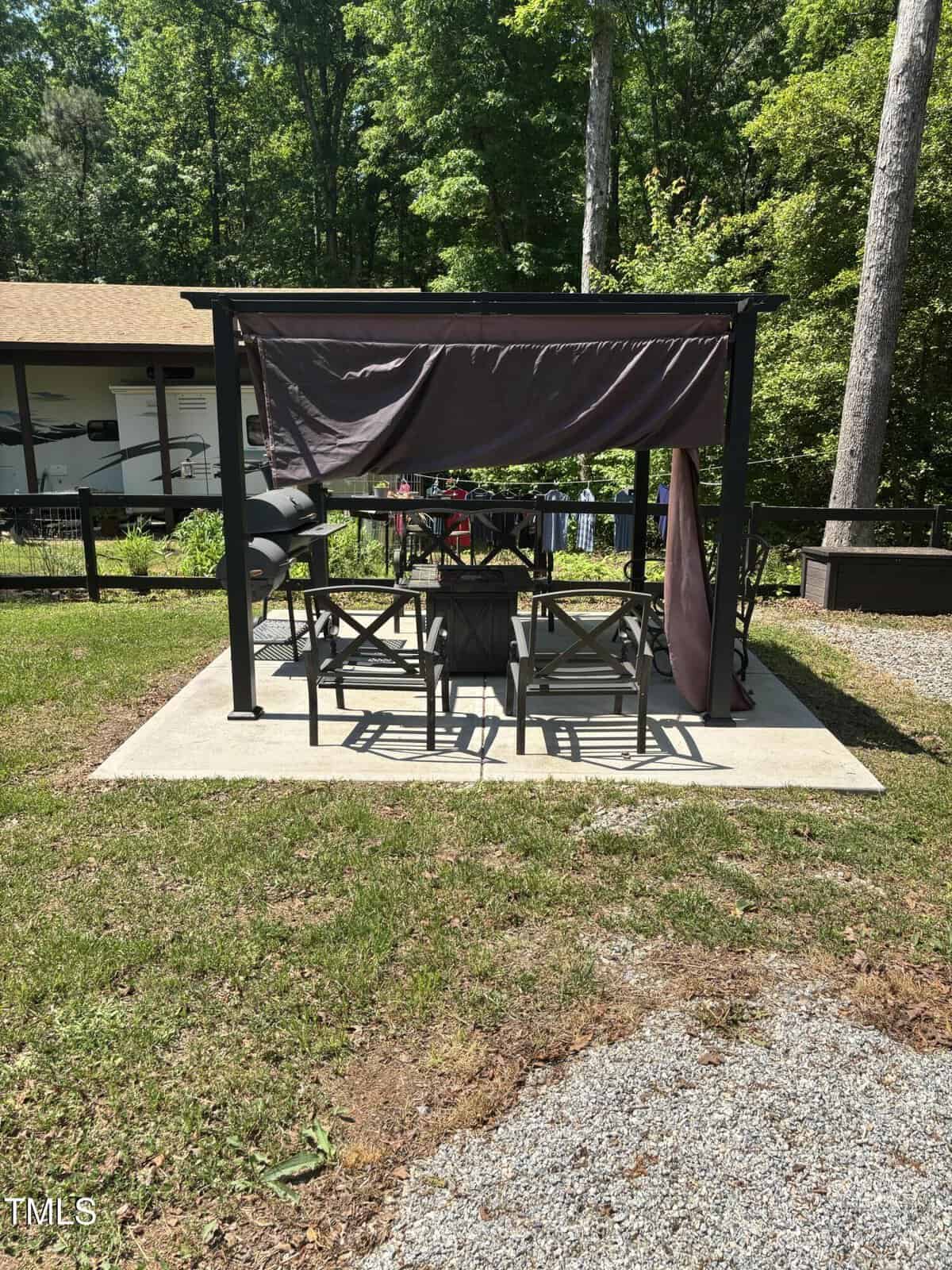 A black metal gazebo with a partially draped brown canopy covers a patio set with chairs and a grill, set on a concrete slab in a grassy yard bordered by trees and a wooden fence.