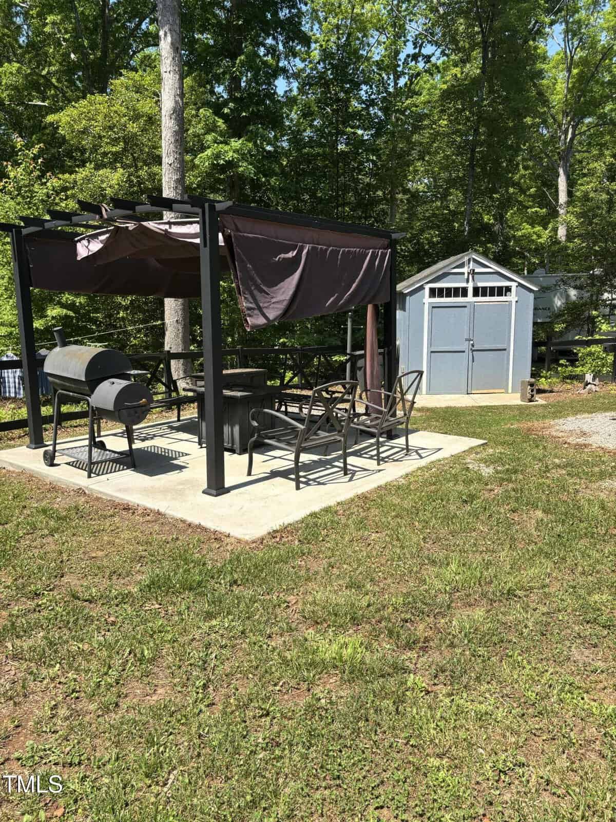 A black metal pergola with chairs and a grill sits on a concrete pad in a grassy yard. A blue-gray storage shed with white trim stands in the background, surrounded by green trees under a clear sky.