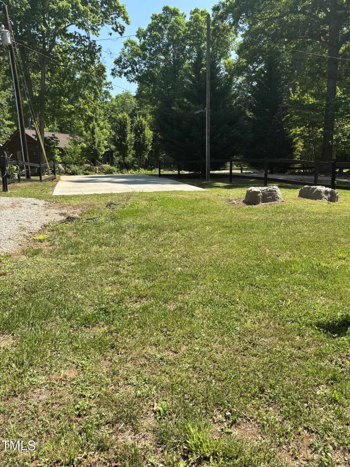A flat concrete slab sits in a grassy yard bordered by trees and rocks. Power lines and a gravel section are visible to the left. A wooden fence runs along the back of the property.