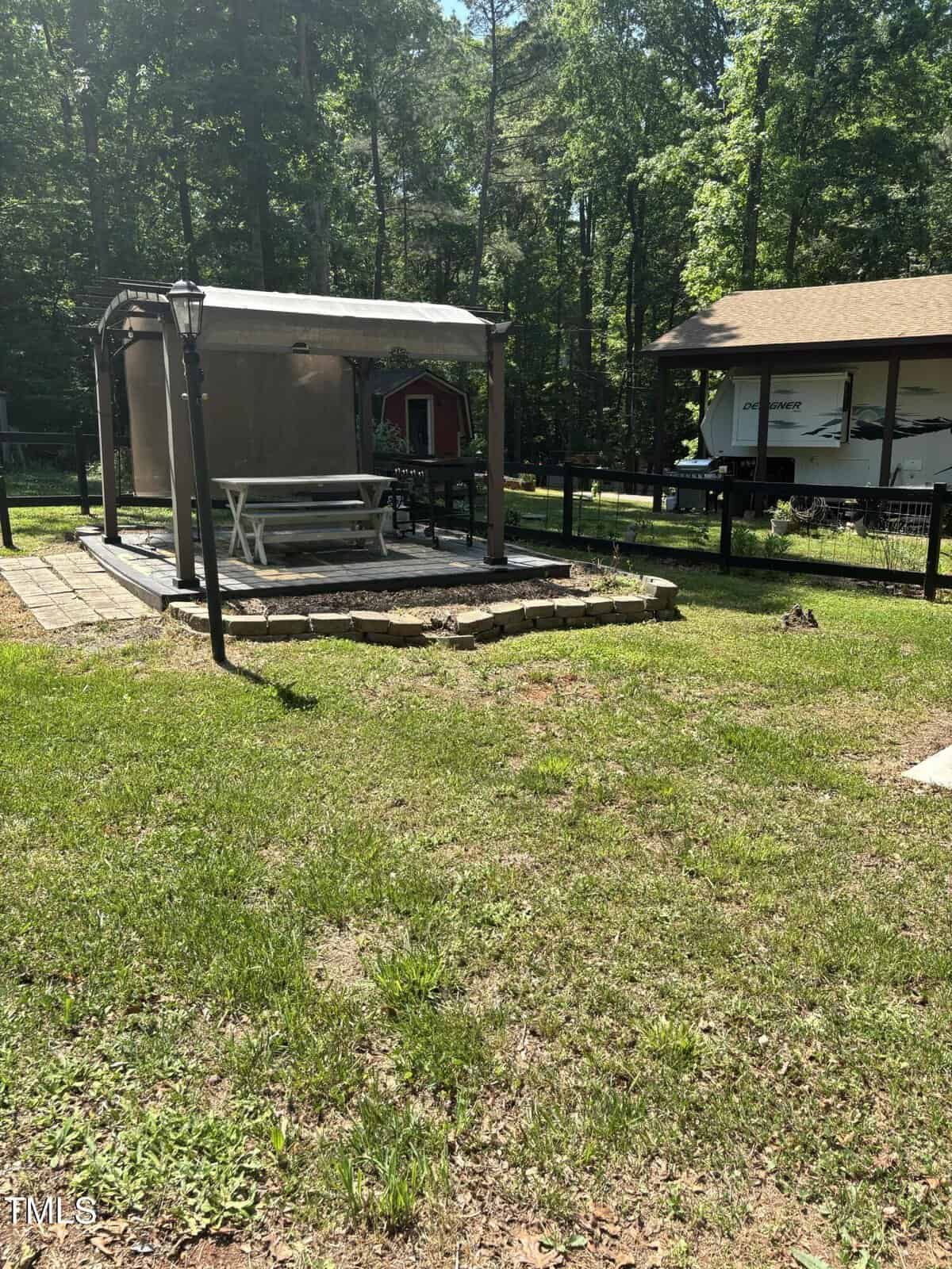 A shaded gazebo covers a white picnic table on a raised stone platform in a grassy backyard. There’s a nearby fence, a lamp post, a red shed, and an RV with trees in the background.