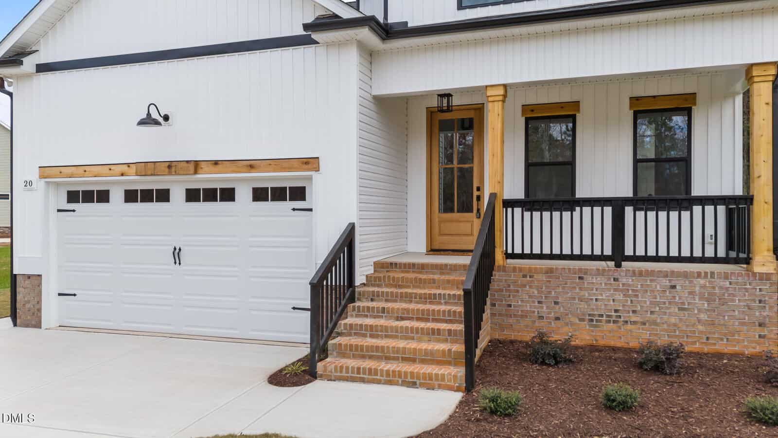 A modern white house exterior with a wooden front door, black railing, brick steps, and a two-car garage with wood accents. There are small shrubs planted along the mulch bed in front.