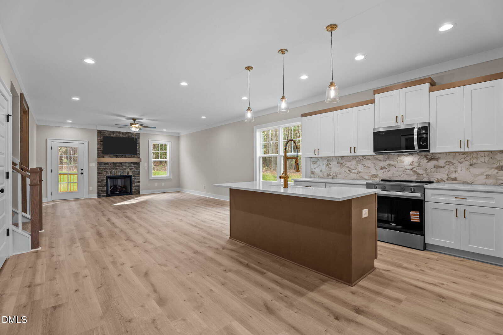 Modern open-concept kitchen and living room with light wood floors, white cabinets, a brown island, pendant lighting, and a stone fireplace. Large windows provide natural light and a view of greenery outside.
