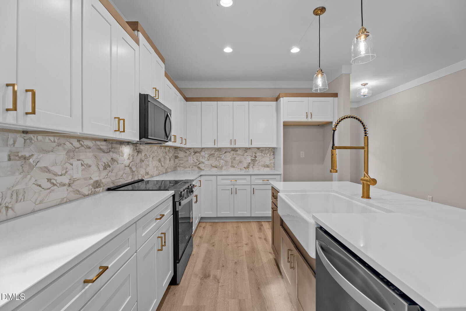 Modern kitchen with white cabinets, gold handles, marble backsplash, stainless steel appliances, a farmhouse sink with a gold faucet, pendant lights, and light wood flooring. The space is clean and well-lit.