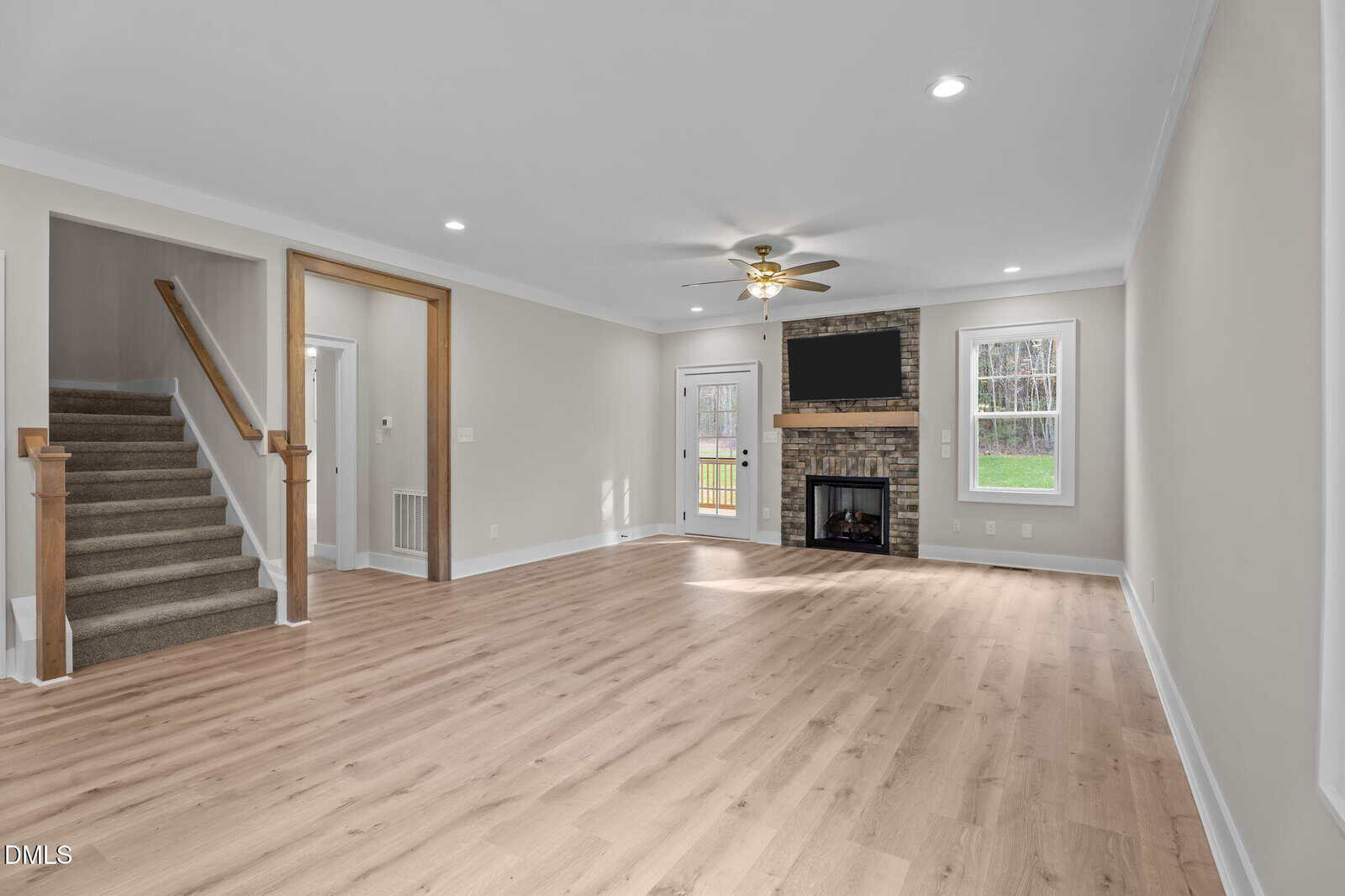 Spacious living room with light wood flooring, beige walls, a stone fireplace with a TV above it, two windows, a ceiling fan, a door to the outside, and a carpeted staircase to the left.