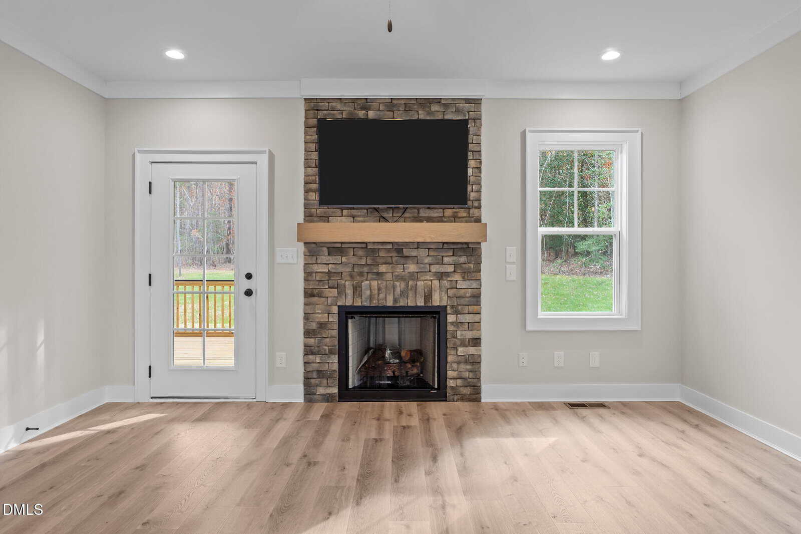 Modern living room with light wood floors, a brick fireplace with a wooden mantel, a TV mounted above the mantel, a glass door on the left, and a window on the right. Walls are painted light beige.